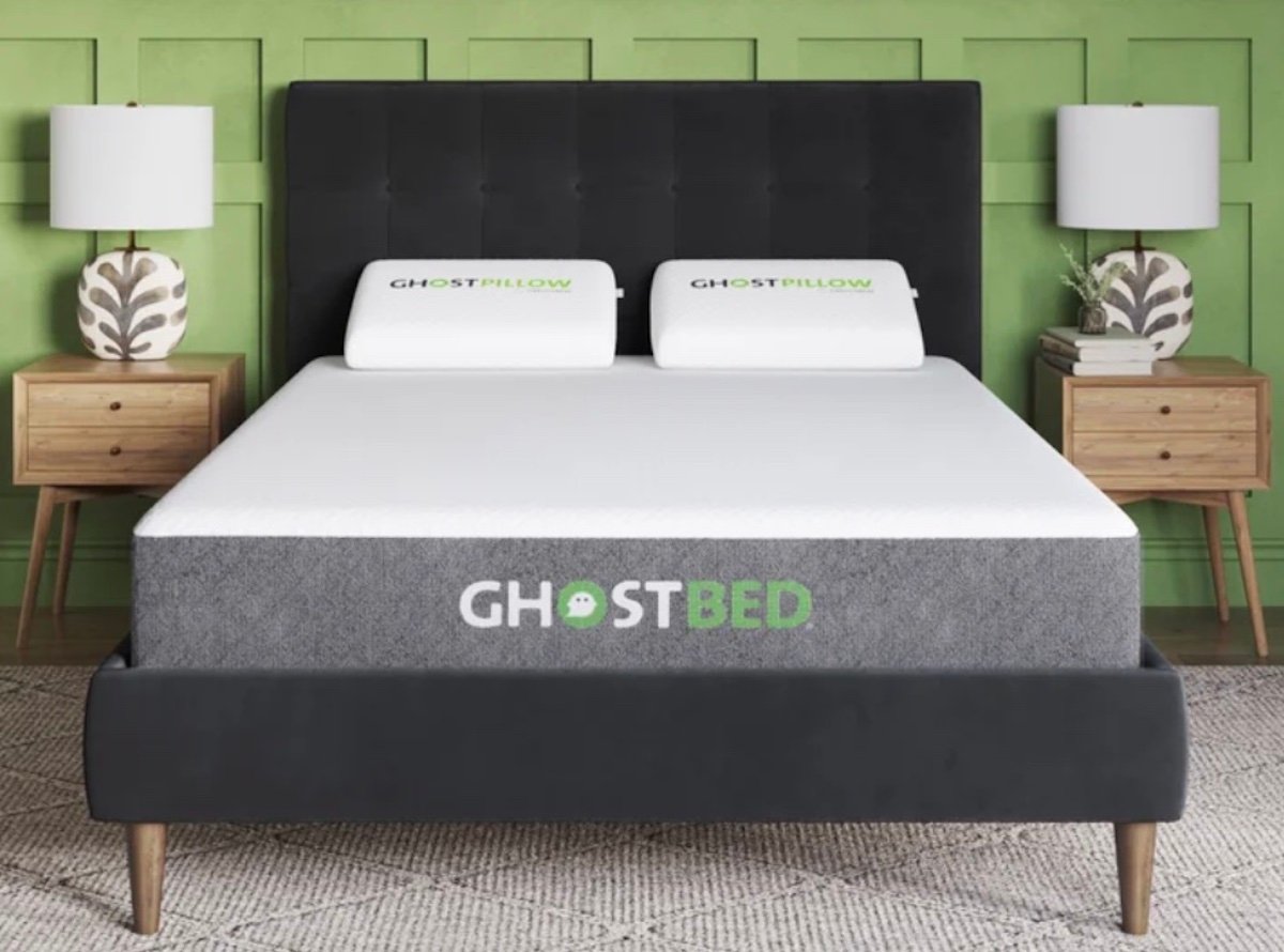 A modern bedroom with a black upholstered bed frame, two GhostPillow pillows, and a GhostBed mattress—styled to inspire comfort like the exclusive Costco deals for seniors. Wooden nightstands with white lamps sit against a calming green wall.