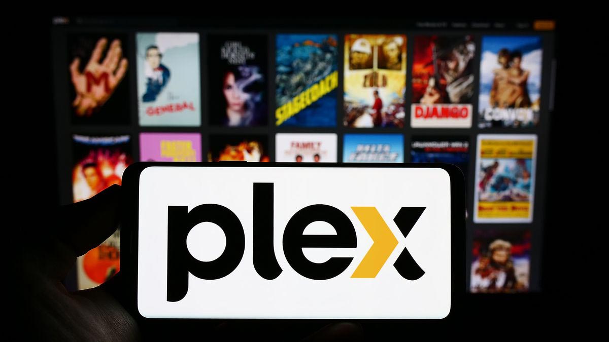A hand holds a smartphone displaying the Plex logo in front of a blurred TV screen filled with colorful movie and TV show posters, highlighting Plex as one of the popular free streaming services available today.