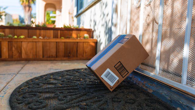 A cardboard package rests at an angle against a white metal door on a doormat outside a house, with a wooden planter and paved walkway visible in the background.