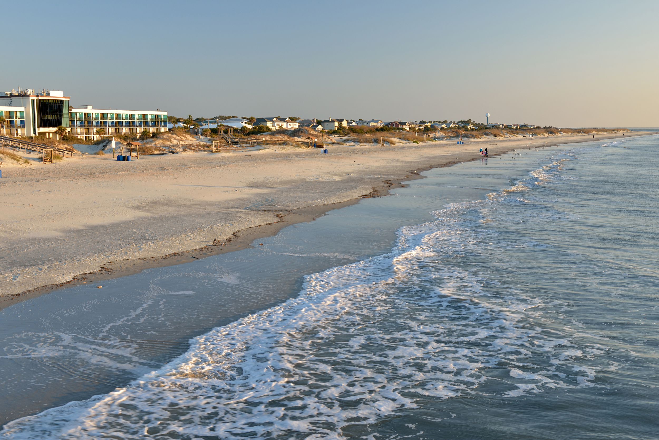 Beach of Tybee Island, Georgia during sunrise, a few people walking along beach, a hotel and homes in the background