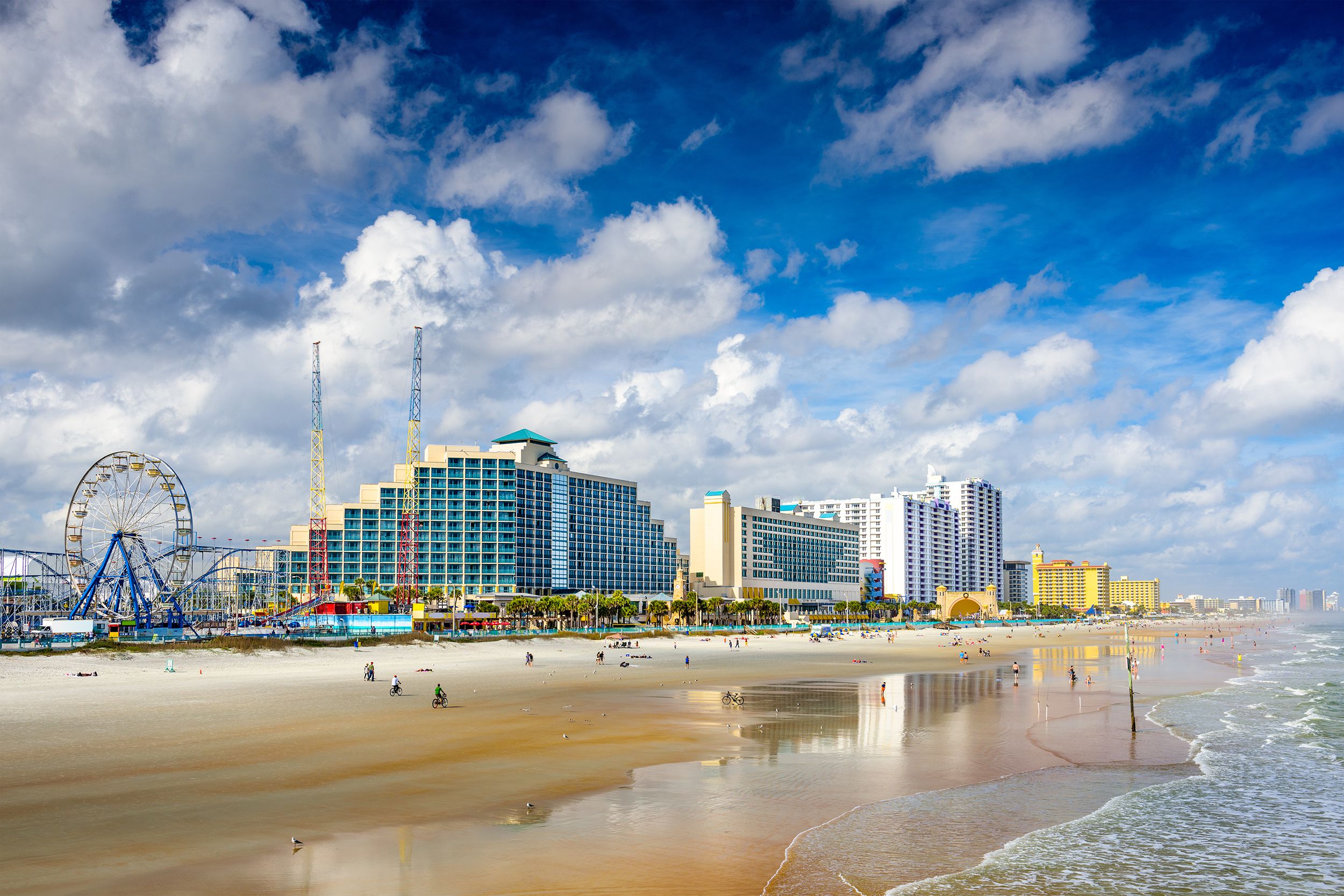 Beach with ocean in the foreground of Daytona Beach, Florida, hotels, a Ferris wheel, and buildings line the background, an expansive blue sky with white and grey clouds