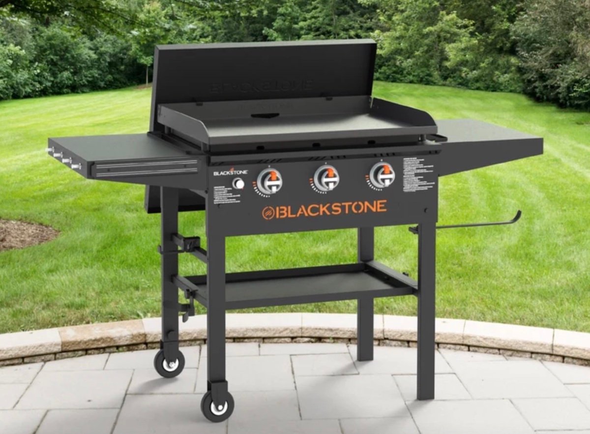 A Blackstone outdoor gas griddle—one of the best Costco finds—with three control knobs, two side shelves, and four wheels is shown on a stone patio with green grass and trees in the background.