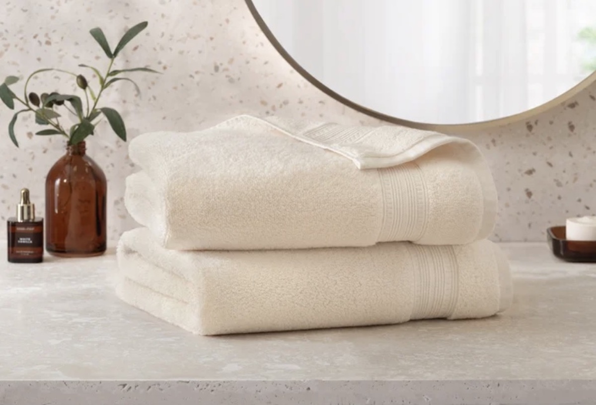 Two neatly folded cream-colored towels—one of the best Costco finds—are stacked on a bathroom countertop. In the background, a brown glass bottle with a plant, a mirror, and a small dish create a clean, minimalistic setting.