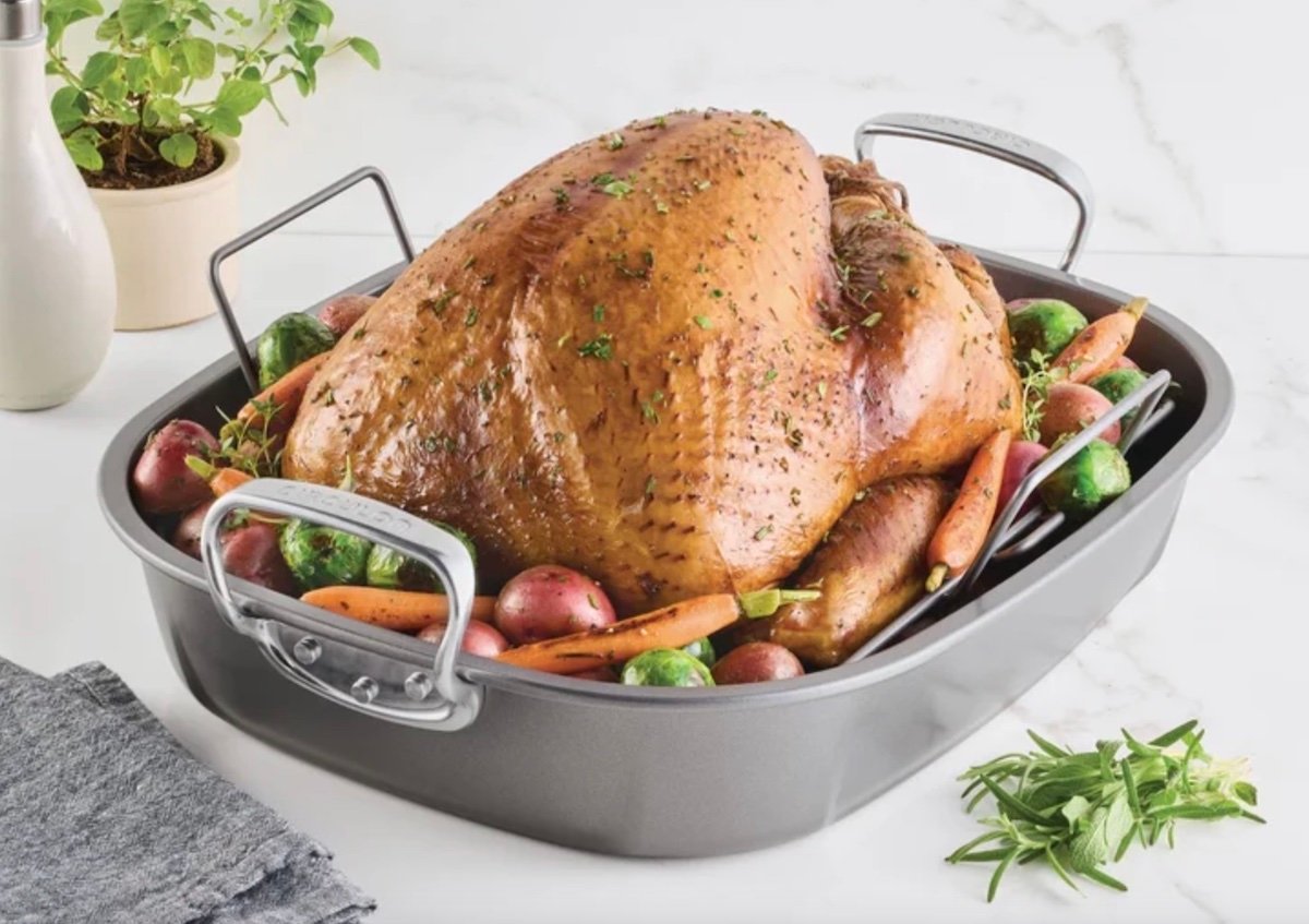 A roasted turkey garnished with herbs sits in a roasting pan surrounded by carrots, Brussels sprouts, and red potatoes on a marble countertop—one of the best Costco finds for an impressive holiday feast. Fresh herbs and a towel are nearby.