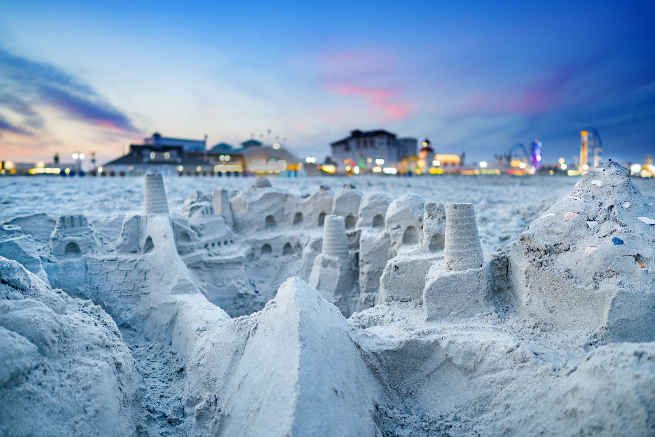 Closeup of an intricate sand castle in the foreground, Ocean City, New Jersey, rest of beach with boardwalk and buildings with lights in the background during sunset, a blue sky with dark blues, pinks, and purples