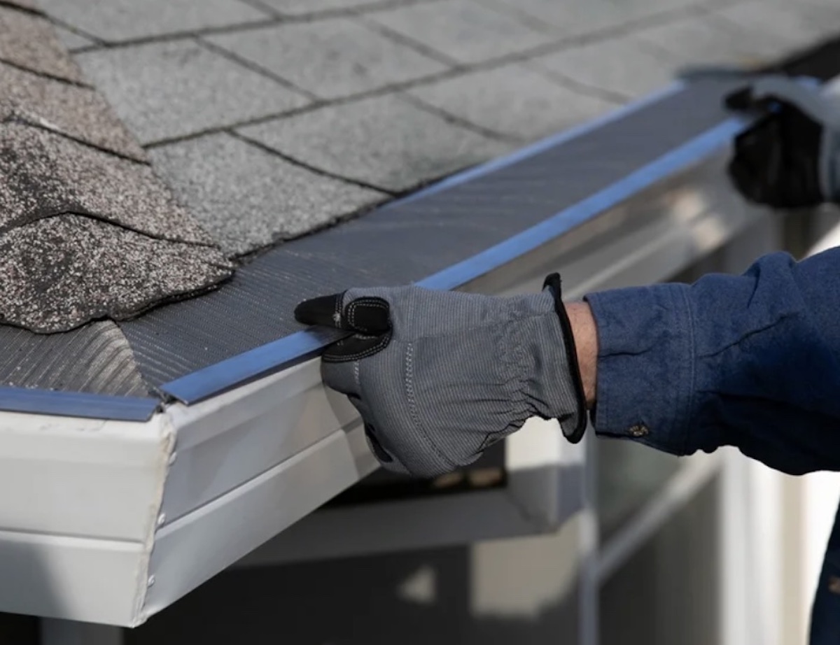 A person wearing gray gloves and a blue shirt installs a mesh gutter guard—one of the best Costco finds—along the edge of a shingled roof, securing it to keep debris out of the house's white rain gutter.