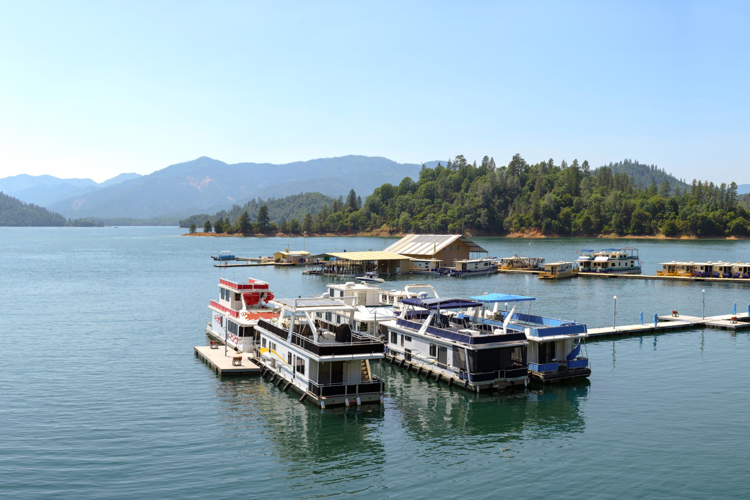 A few houseboats docked in a marina of Shasta Lake, California, evergreen trees and mountains in the background on a sunny day