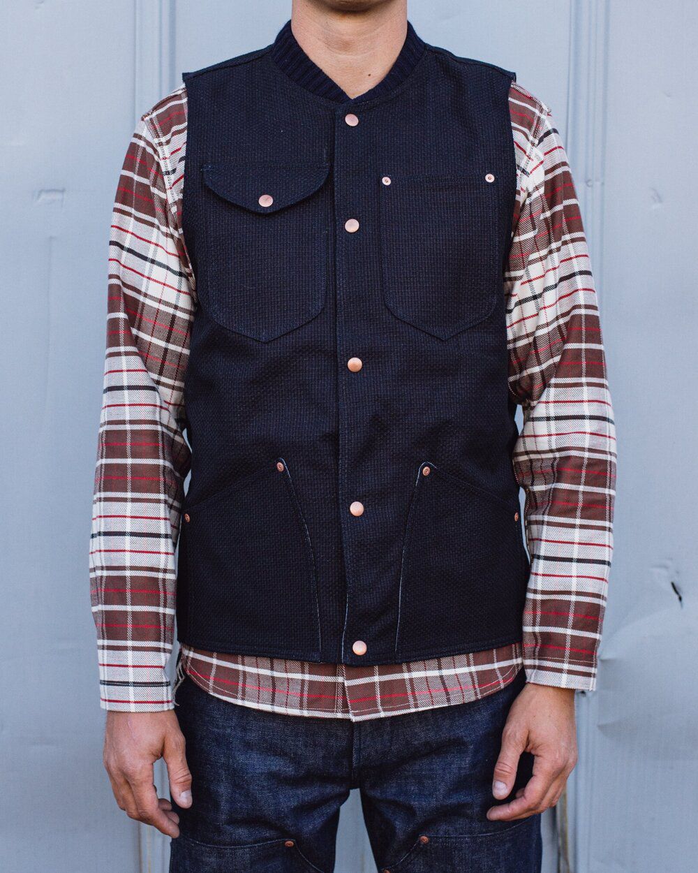 Model wearing long sleeve checkered button down shirt, buttoned dark denim vest and dark jeans.Grease Point Workwear