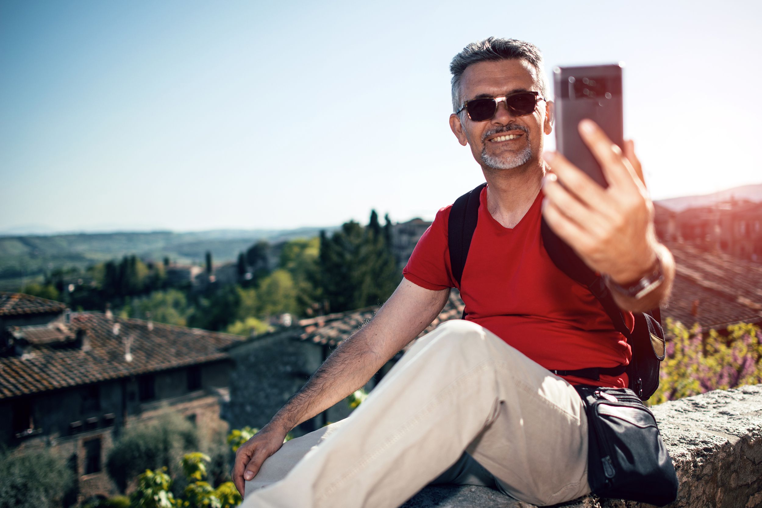 Happy senior bearded man smiling while taking selfie photo with smartphone and wearing eyeglasses outdoors in Florence, Italy