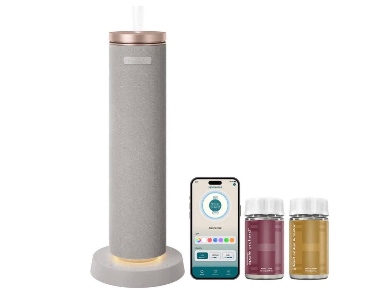 A tall, cylindrical smart drink dispenser with a sleek gray and rose gold design sits on a base—one of the best Costco finds—accompanied by two drink pods and a smartphone displaying the device’s app interface with drink options.