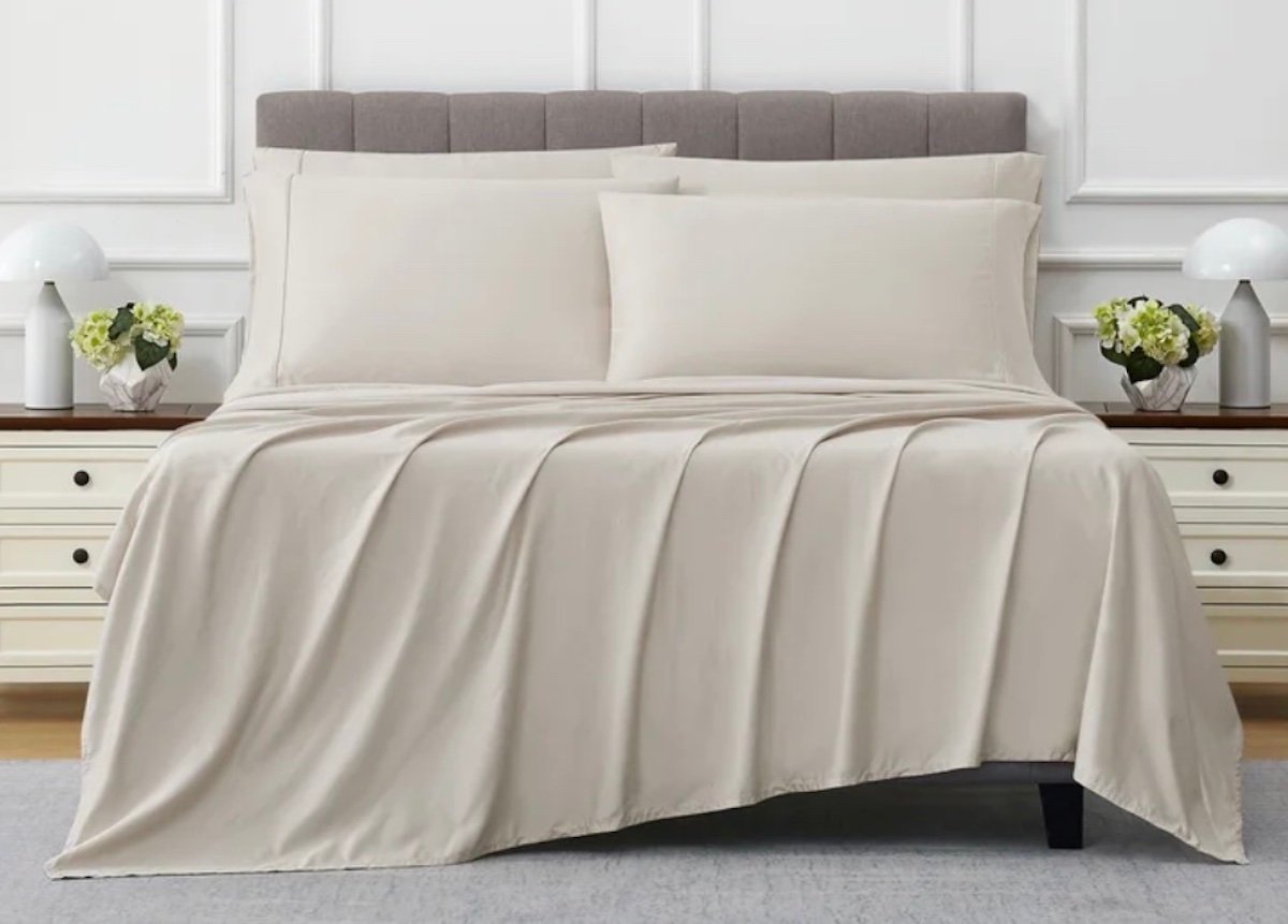 A neatly made bed with light beige bedding, two large pillows, and a padded gray headboard—one of the best Costco finds. Nightstands with white lamps and flower arrangements sit on each side, set against a white paneled wall.