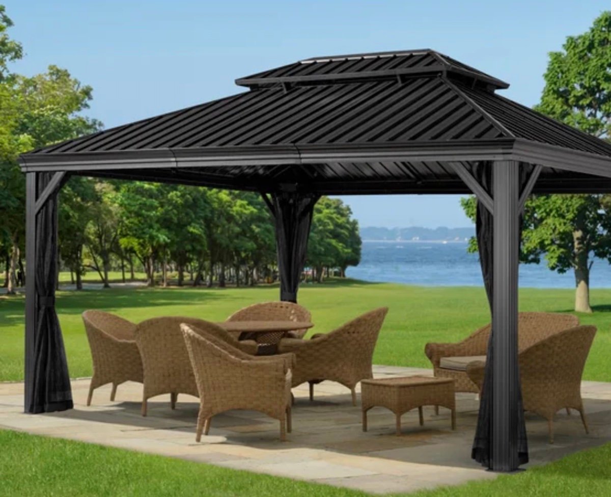 A black metal gazebo—one of the best Costco finds—with wicker chairs and tables stands on a stone patio in a grassy park near a lake, framed by trees and blue sky in the background.