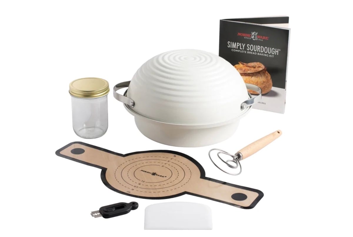 A sourdough bread baking kit—one of the best Costco finds—includes a lidded baking bowl, recipe book, glass jar, dough whisk, scraper, silicone mat, and thermometer, all arranged on a white background.