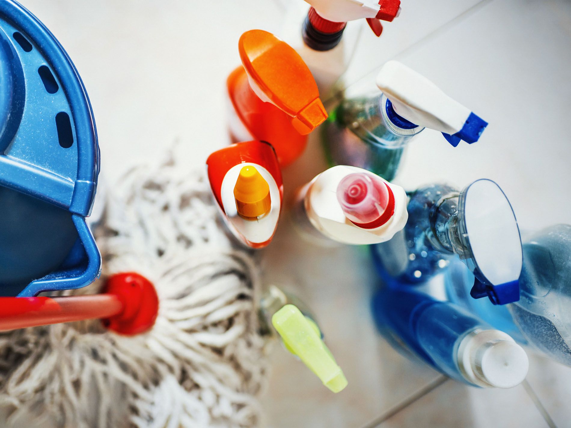 Closeup top view of unrecognizable home cleaning products with blue bucket and a mop on the side. All products placed on white tiled bathroom floor.