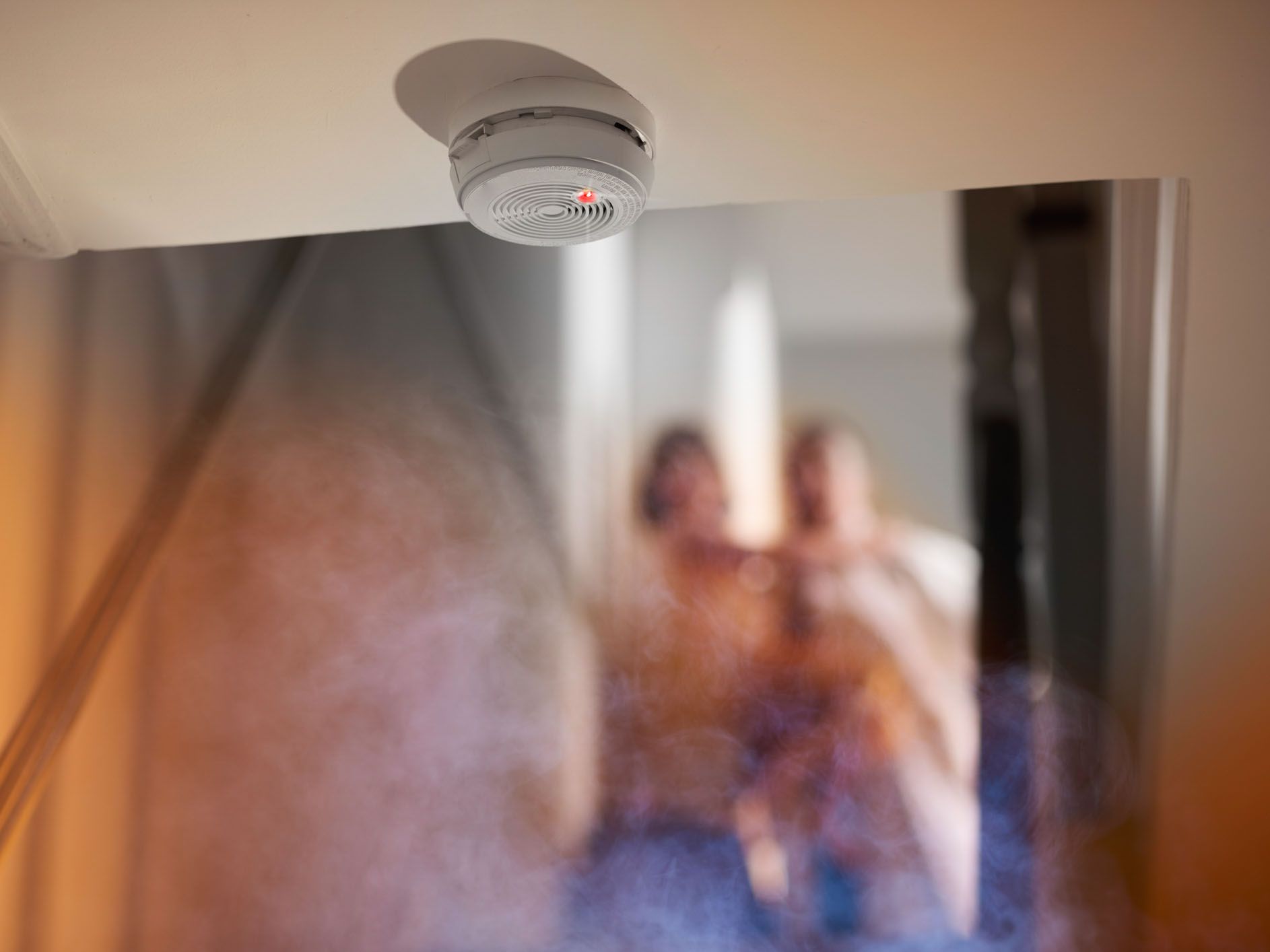 smoke rises up towards a second floor in a two two storey house . Upstairs we can see a parent carrying a small child and running from a bedroom as they have been woken by the smoke alarm which is shown in focus at the edge of the ceiling leading to the s