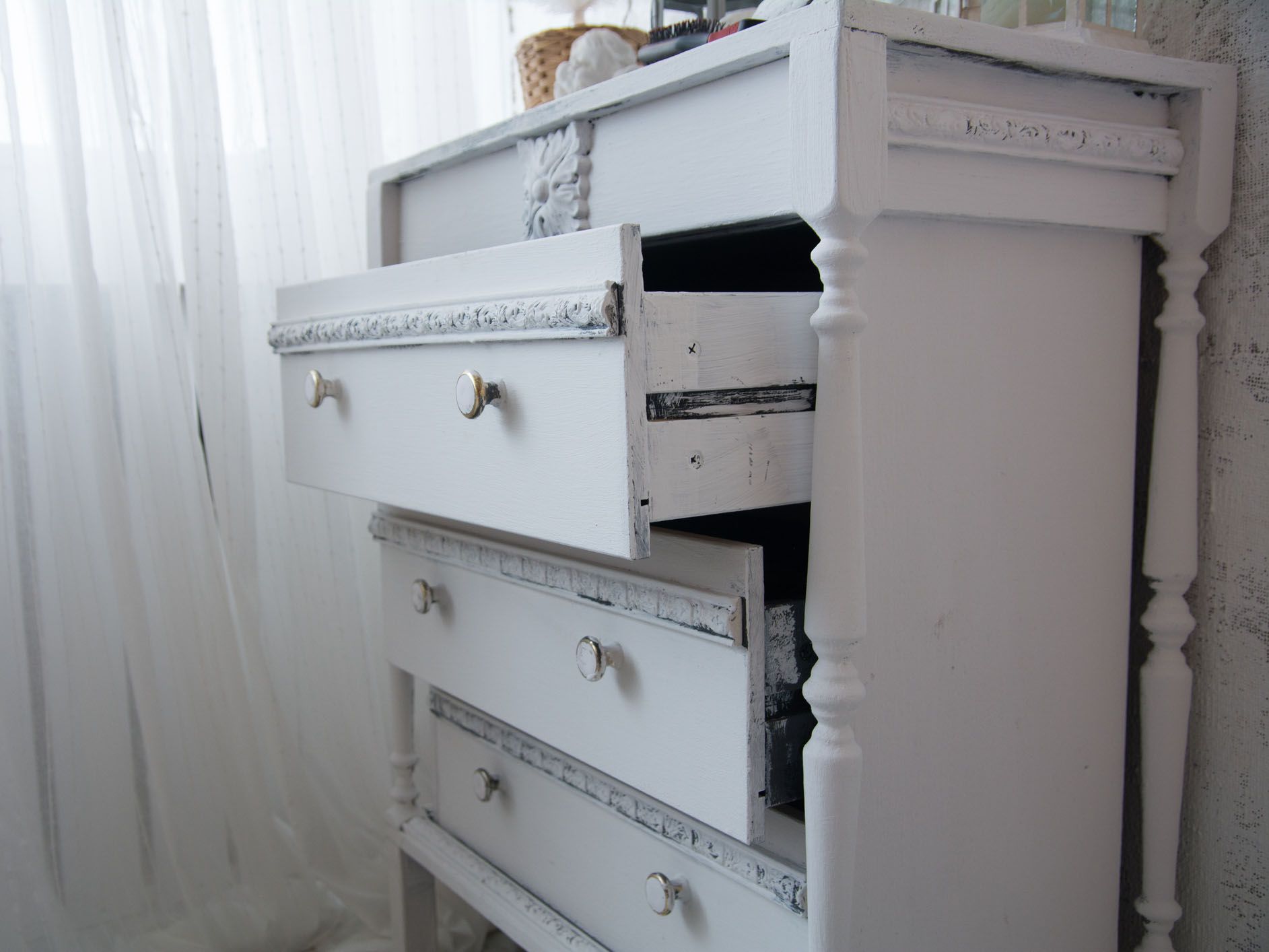 An old painted white ladies chest of drawers with open drawers by the window. Ladies' retro table near the cloth wall