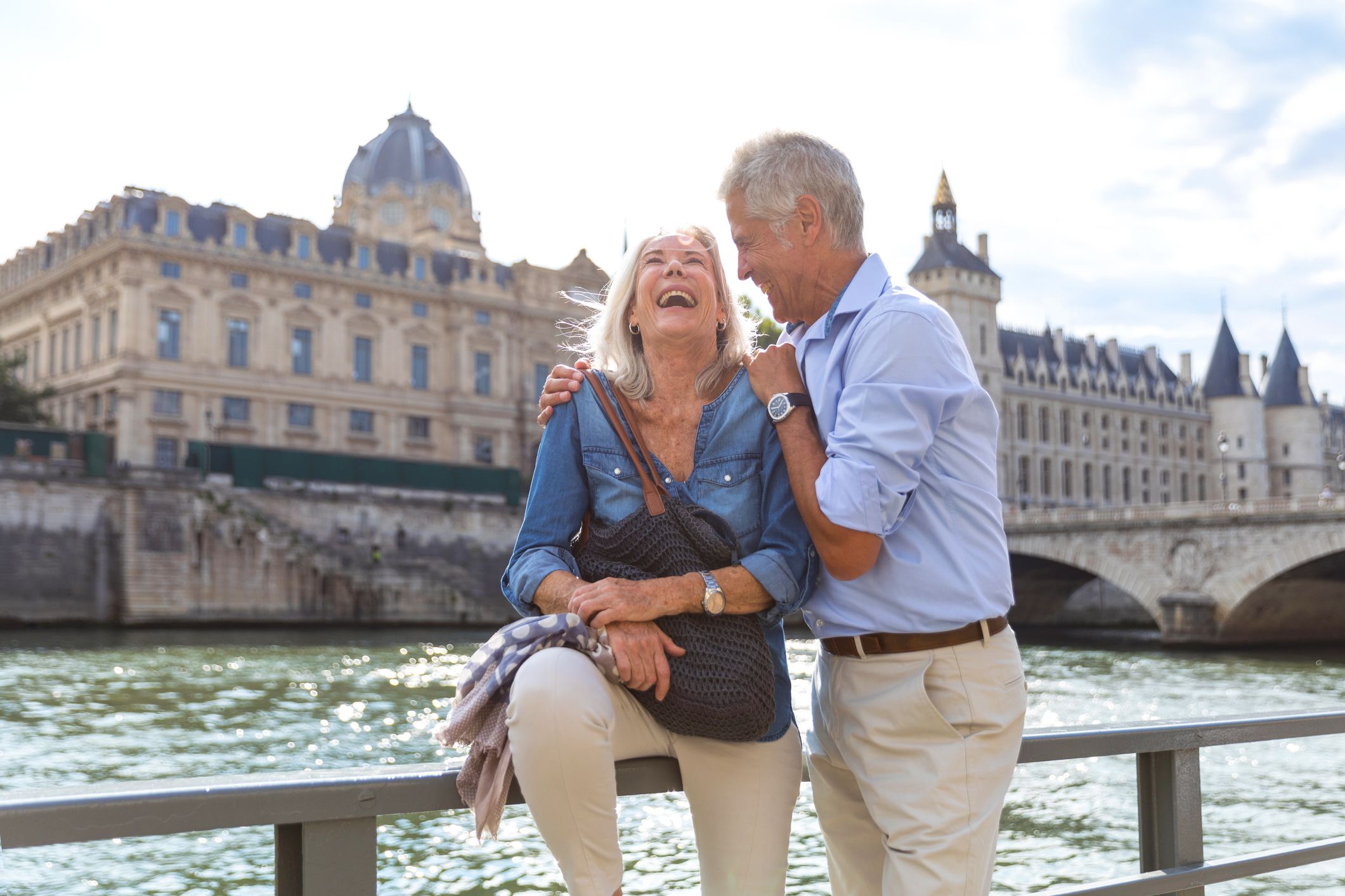 Senior couple relaxing along the Seine River in Paris, woman is laughing while man is looking at her and hugging her, with buildings in the background