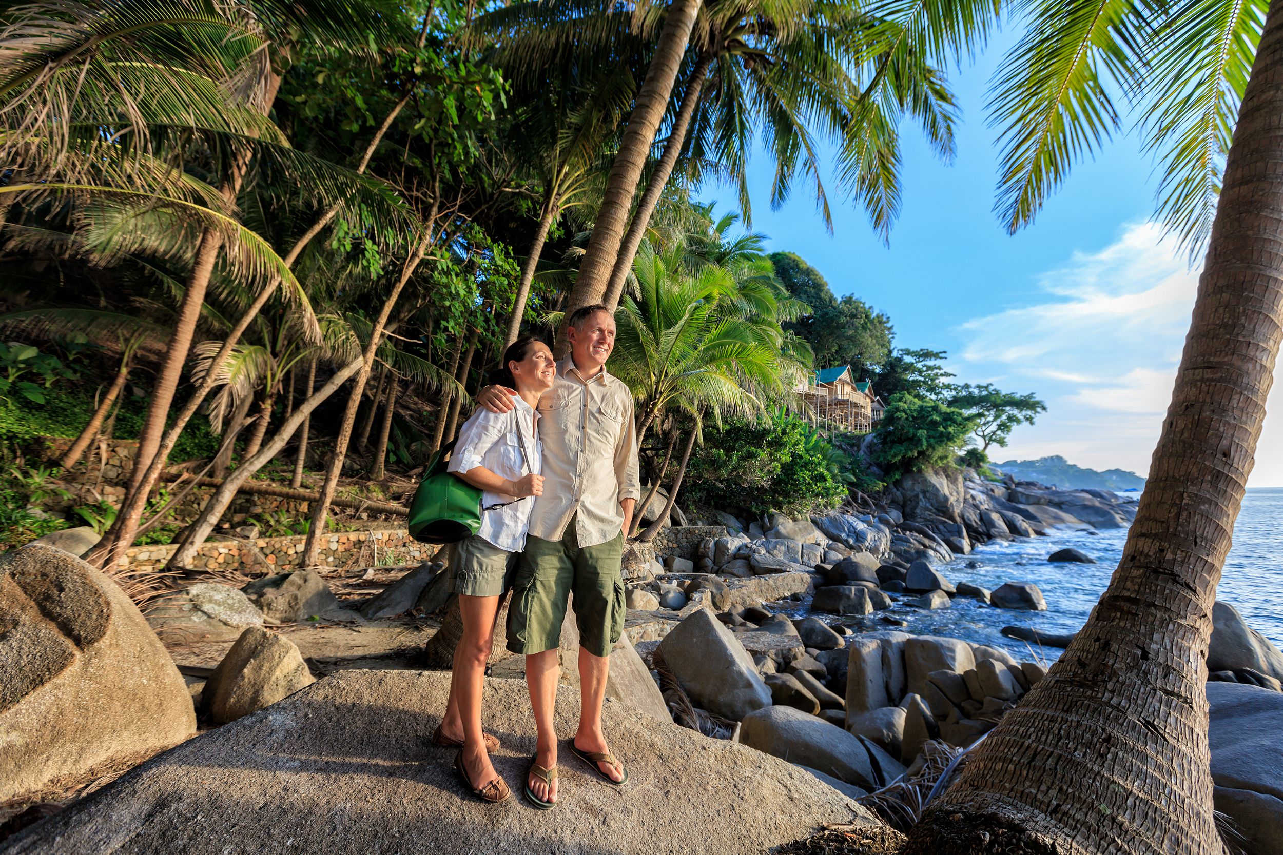Senior couple at romantic tropical beach in Thailand, couple standing on rocks of beach with palm trees overlooking the ocean on a sunny day