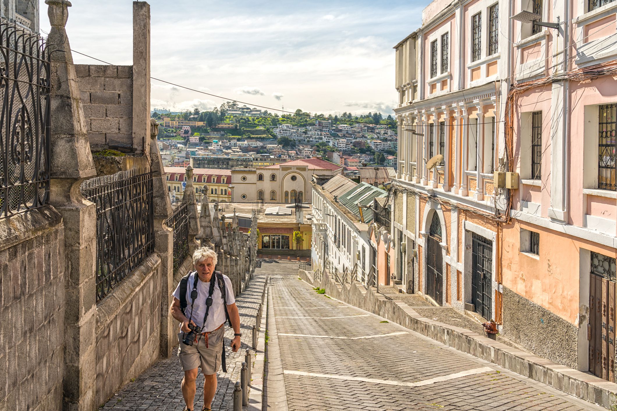 Senior man with camera going up a steep street in old town of Quito, Ecuador, buildings on both sides, city and greenery in the background