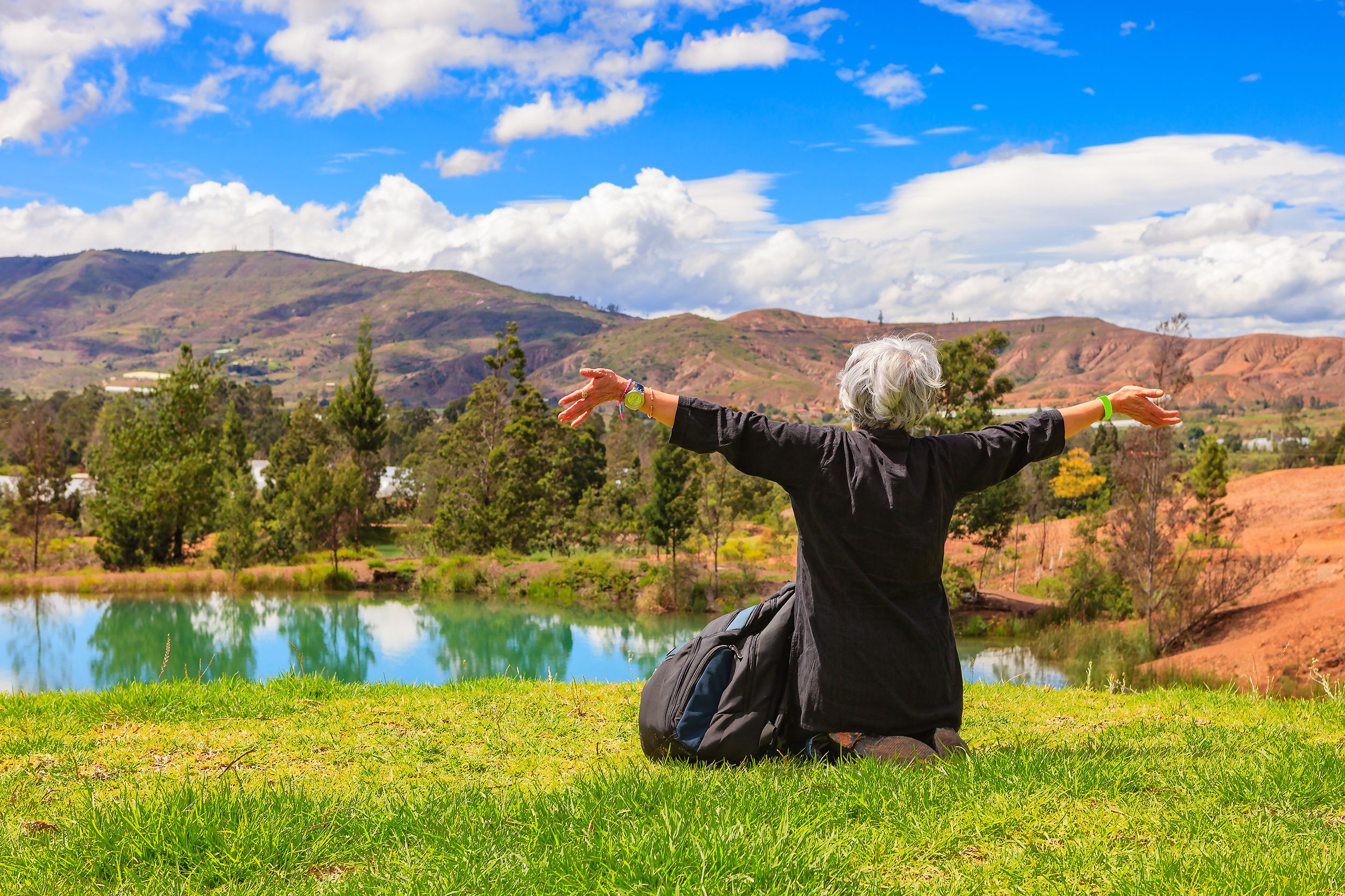 Senior woman enjoying the scenery of the Andes Mountains, Colombia, she is sitting in the grass with her arms stretched out in front of a lake and the mountains and blue sky in the background