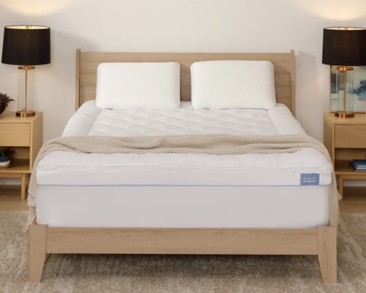 A neatly made bed with a light wood frame, two white pillows, a beige blanket, and a white mattress topper—one of the best Costco finds. Matching nightstands with black lamps and decor complete the room’s cozy, minimalistic style.