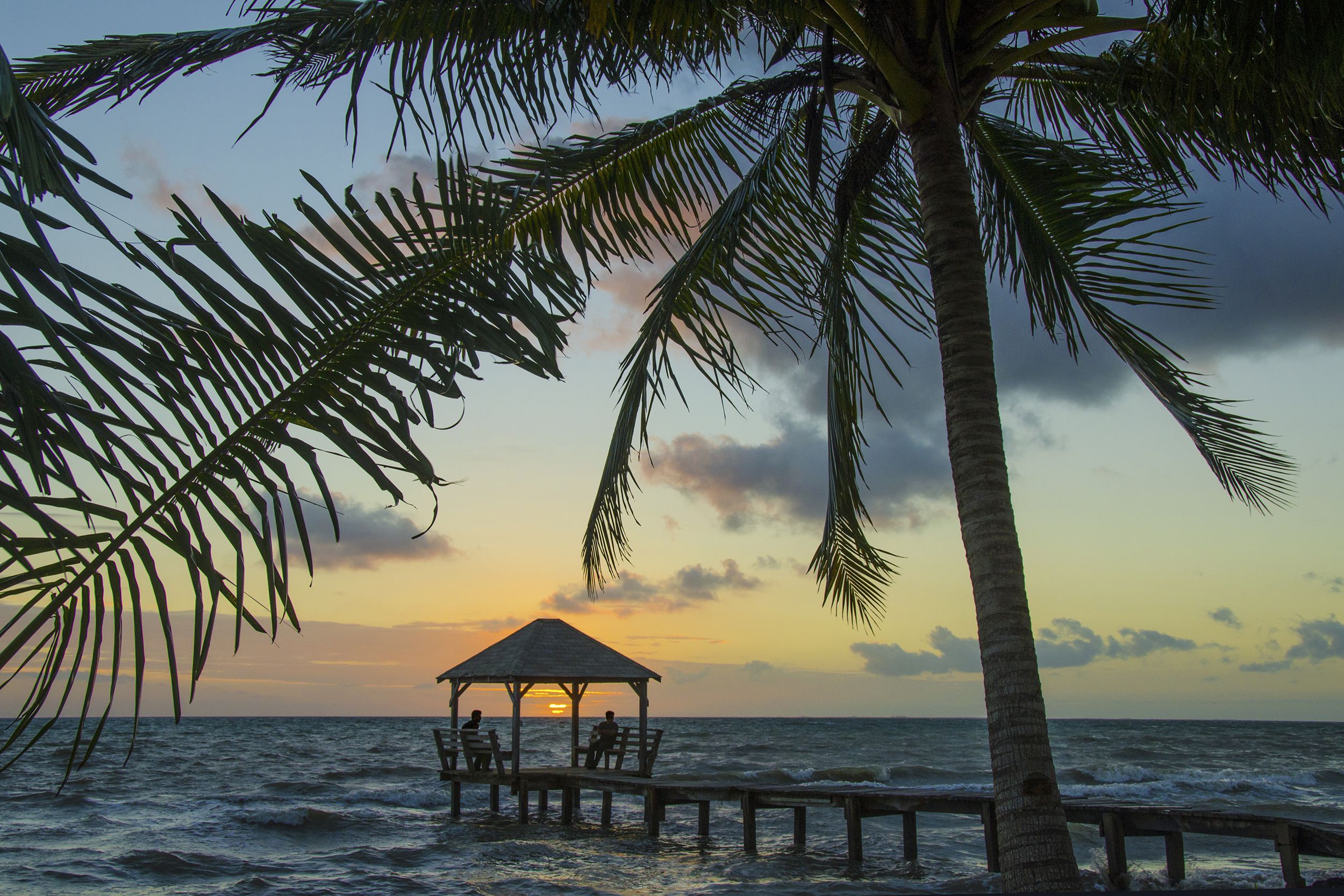 Silhouette of one palm tree in the foreground and two people in the distance watching sunrise from under palapa at end of pier in Placencia, Belize