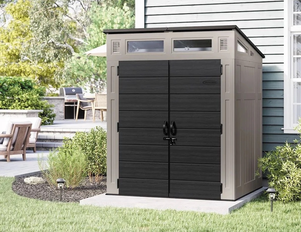 A beige and dark gray outdoor storage shed—one of the best Costco finds—features double doors and small upper windows, perched on a concrete slab next to a house, surrounded by green plants and a patio with chairs in the background.