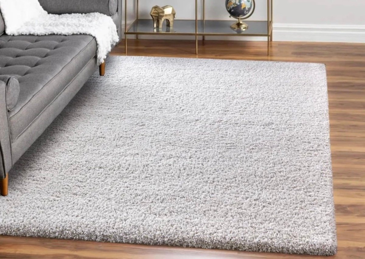 A modern living room with a light gray shag rug on wooden flooring, part of a gray sofa with a white throw on the left, and a gold shelf showcasing decor items—featuring some of the best Costco finds like a globe and elephant figurine—in the background.