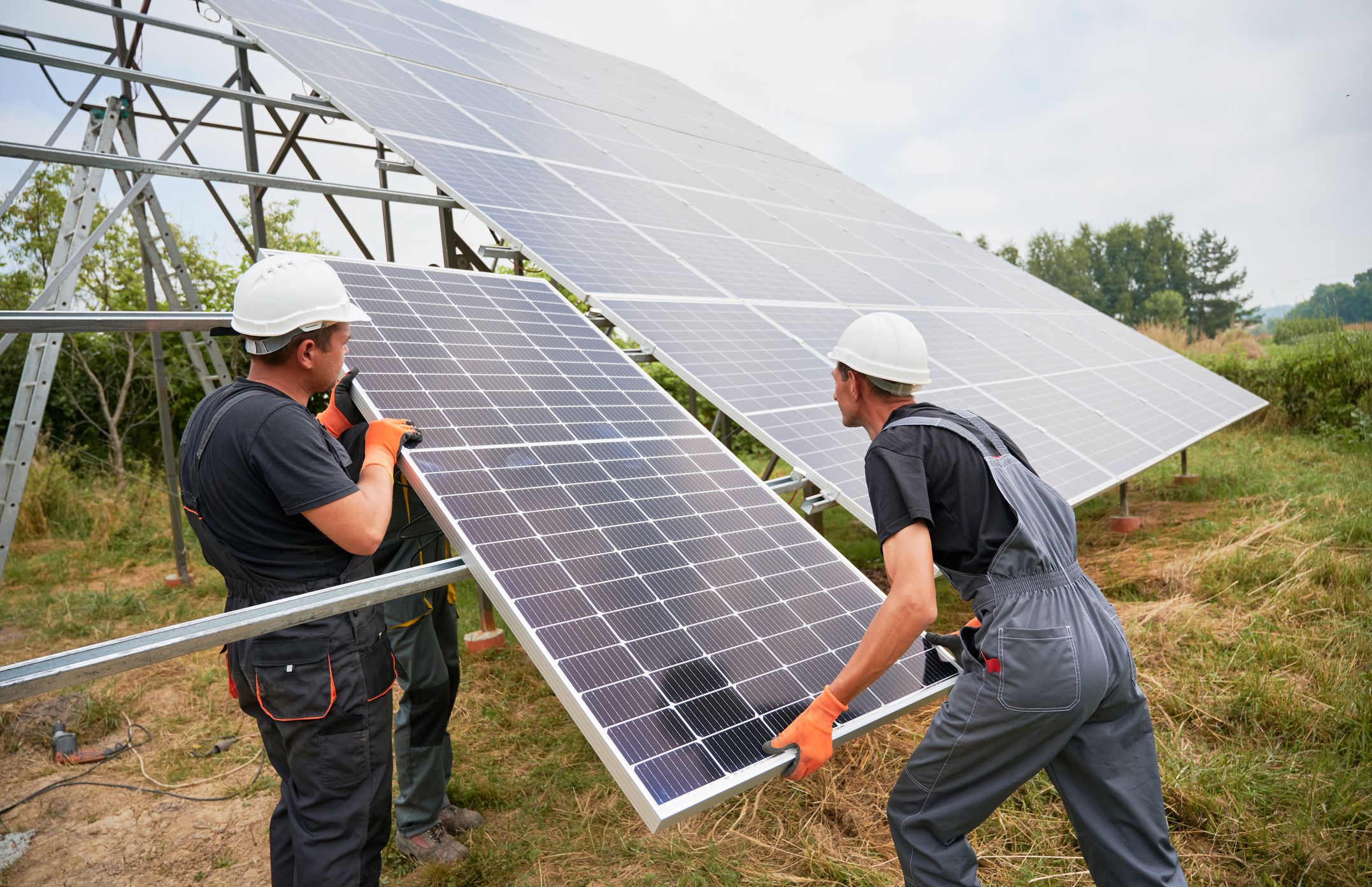 Workers installing solar panel on metal beams in field at daytime. Men wearing workwear and helmets. Ecological energy. Photo-voltaic collection of modules. Array - system of photo-voltaic panels.