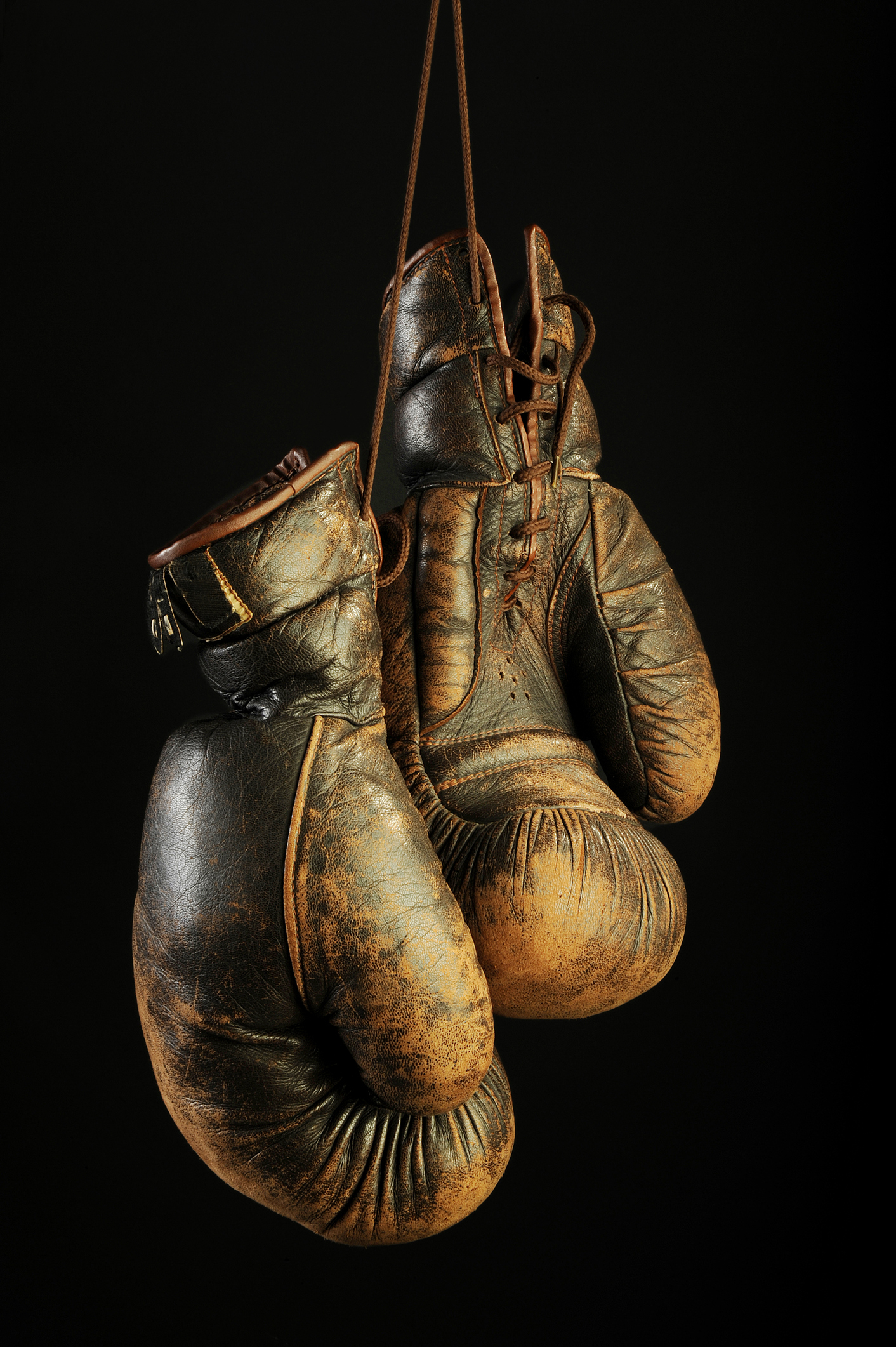 A pair of worn brown vintage boxing gloves hanging from ties shadowed on a black background