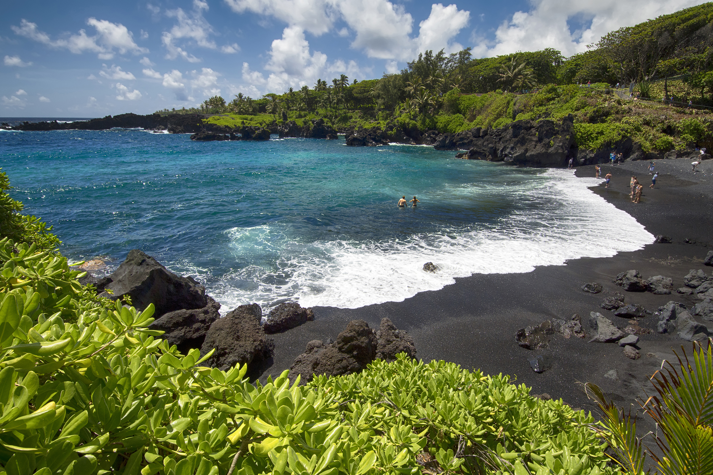 Waianapanapa State Park, Maui, Hawaii, black sand beach in the foreground with lava rocks, surrounded by tropical foliage on a sunny day