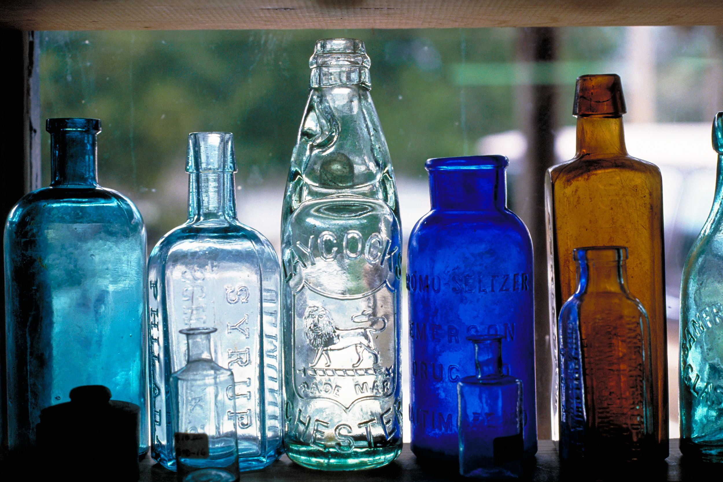 Closeup of clear, aqua, and brown glass bottles in an antique store window seen from inside the store, with a blurred background of outside