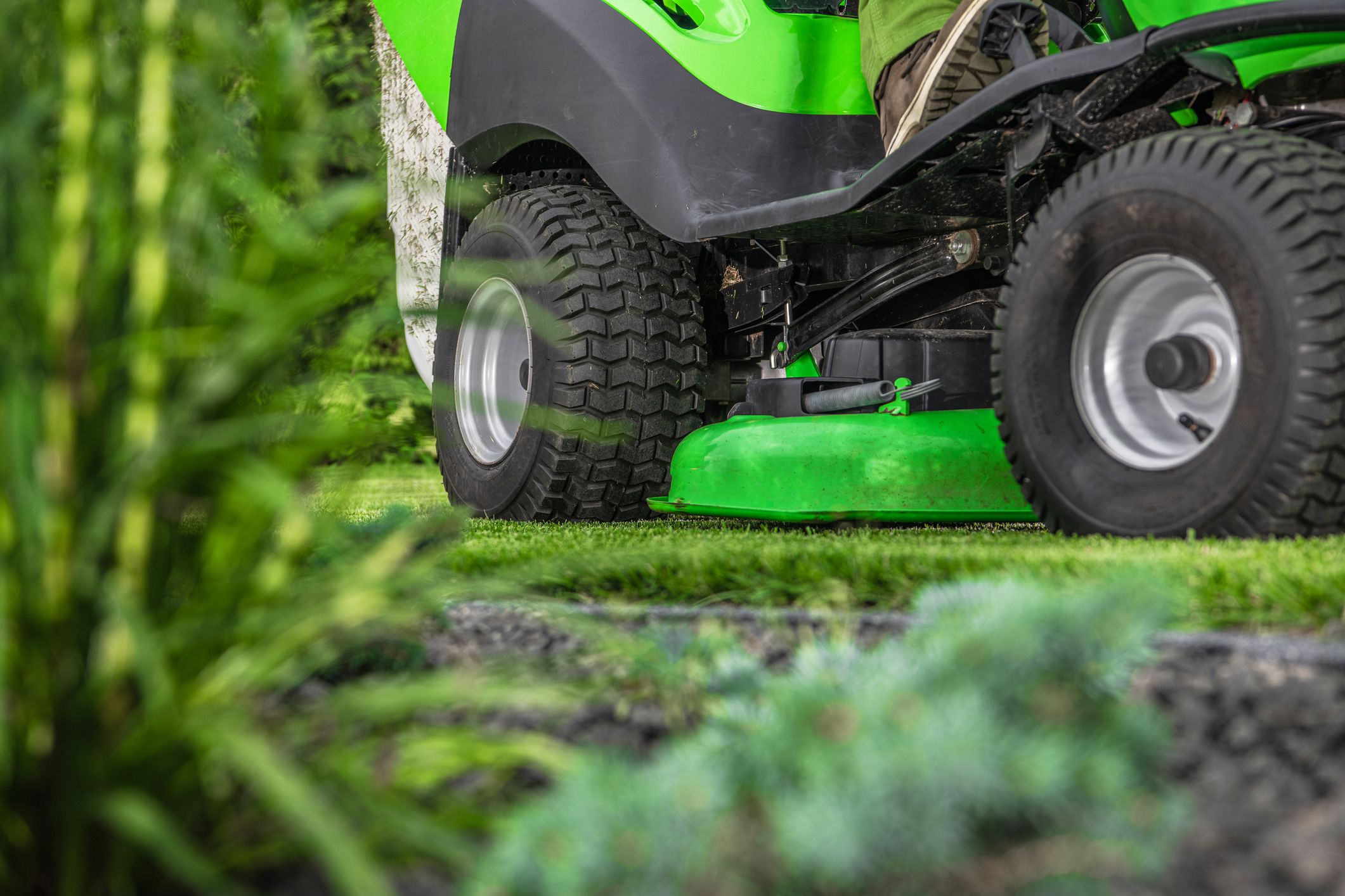 Closeup of green riding lawn mower cutting grass, selective focus, green plants blurred in the foreground