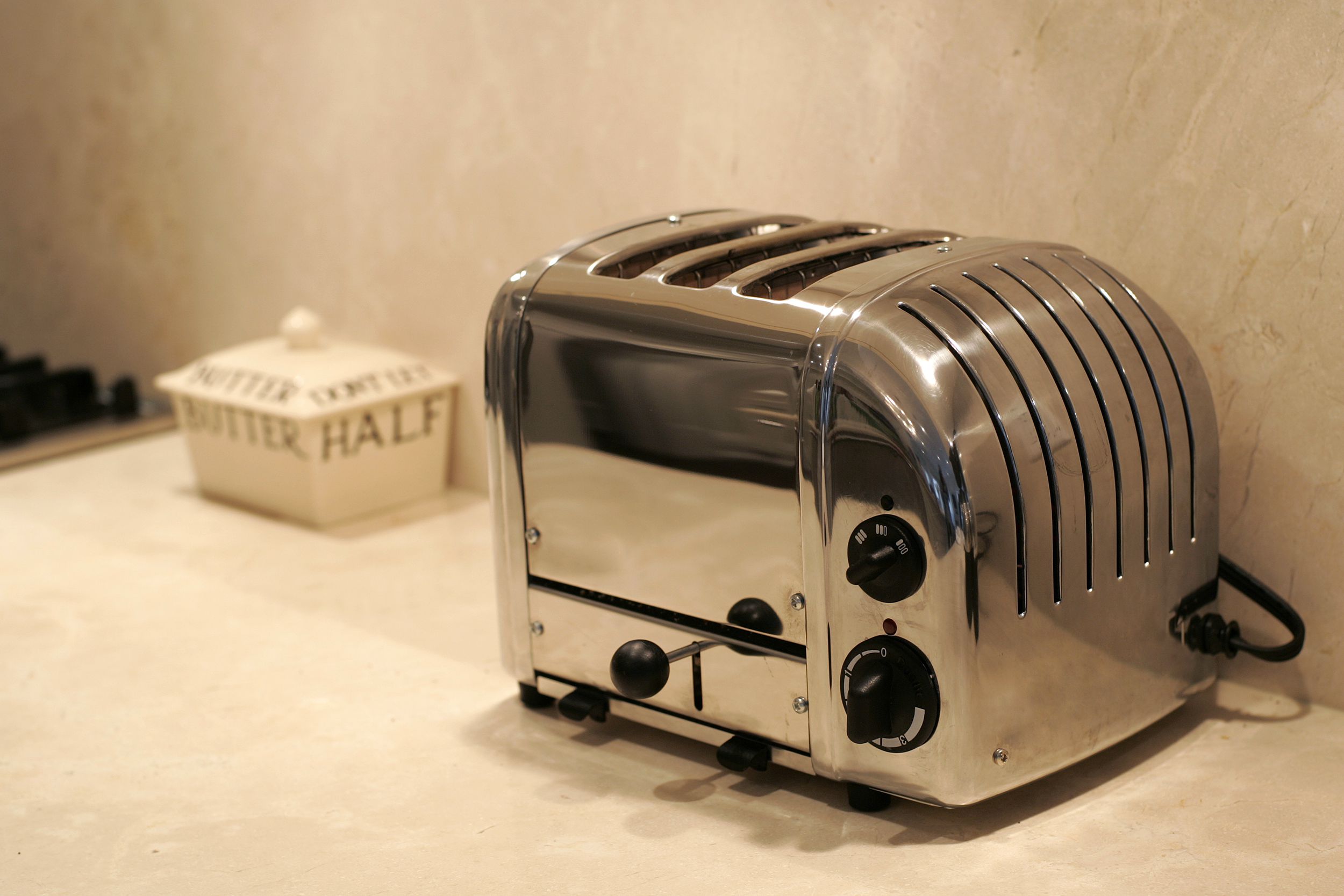 Focus on a chrome vintage toaster on a beige kitchen counter and wall with a beige butter holder and a sliver of a range in the background