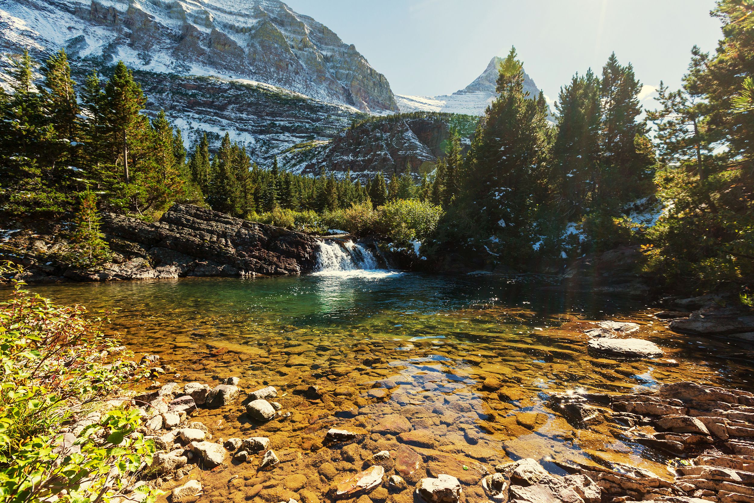 Glacier National Park, Montana, rocks in river in the foreground, evergreen trees line the river, snow on mountains in the background
