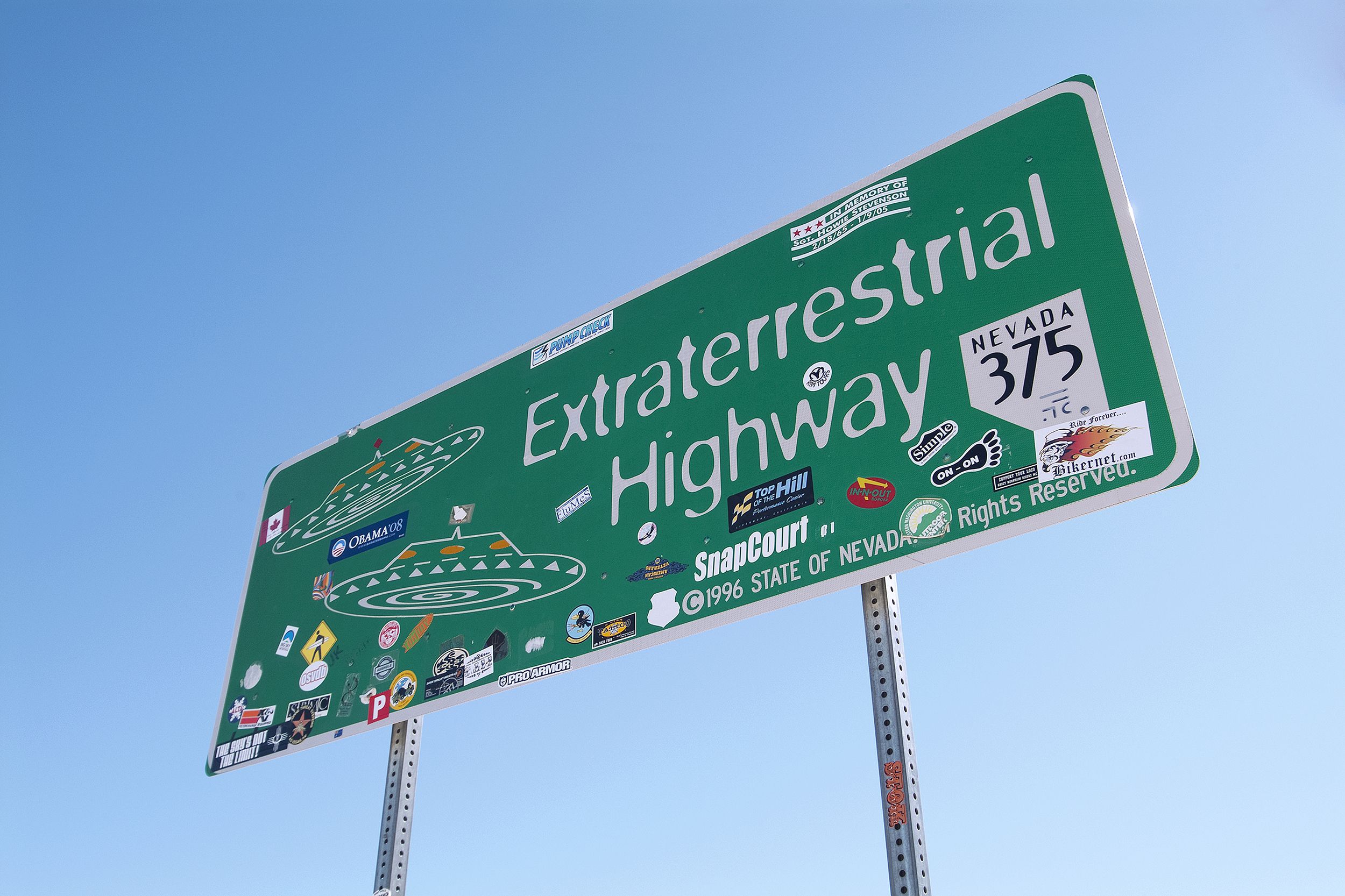 Green highway sign of Extraterrestrial Highway, Nevada, diagonal with stickers, against a clear blue sky