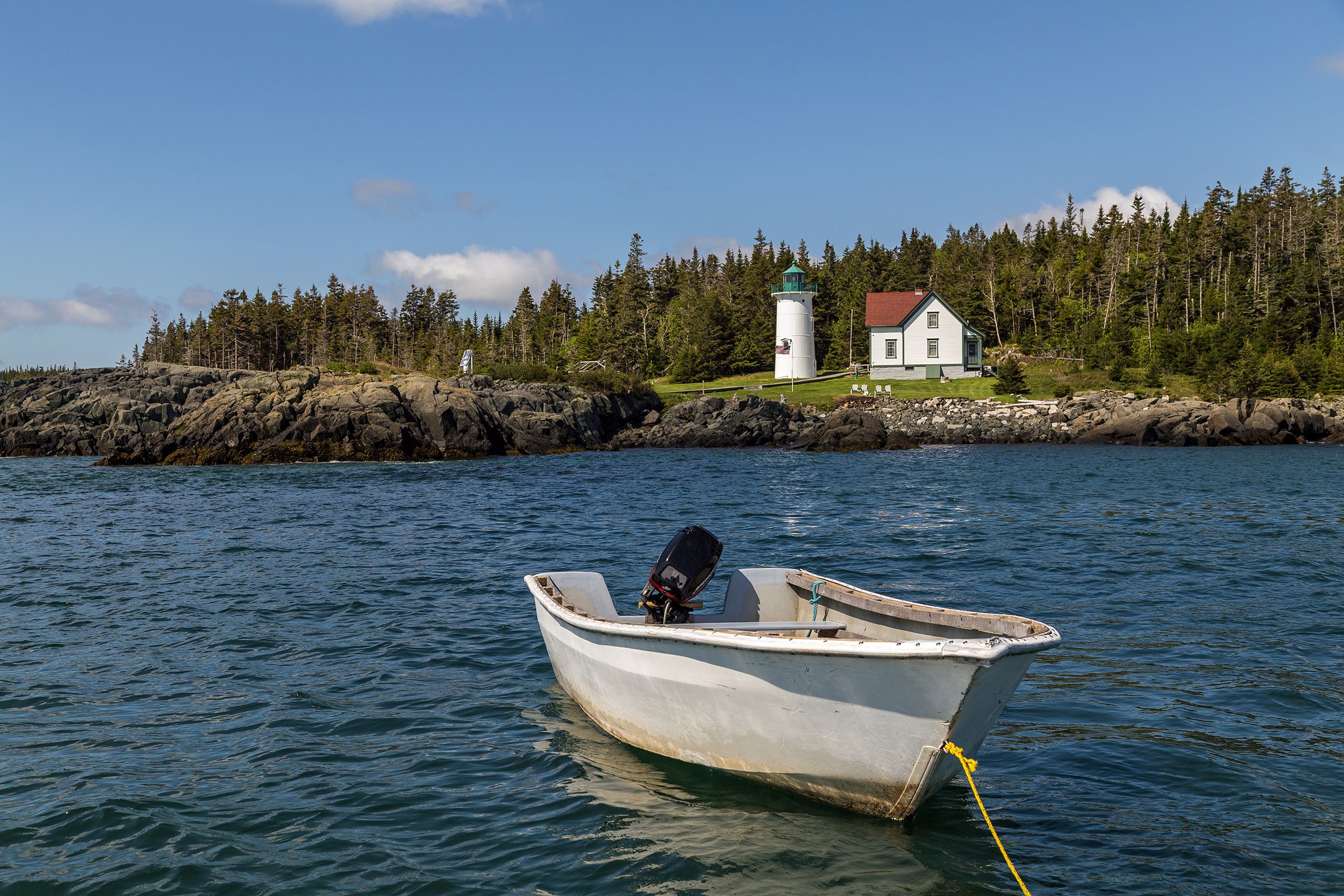 Small white boat in the water in the foreground, Little River Lighthouse, Cutler, Maine, rocky cliffs, evergreen trees on the land on a sunny day