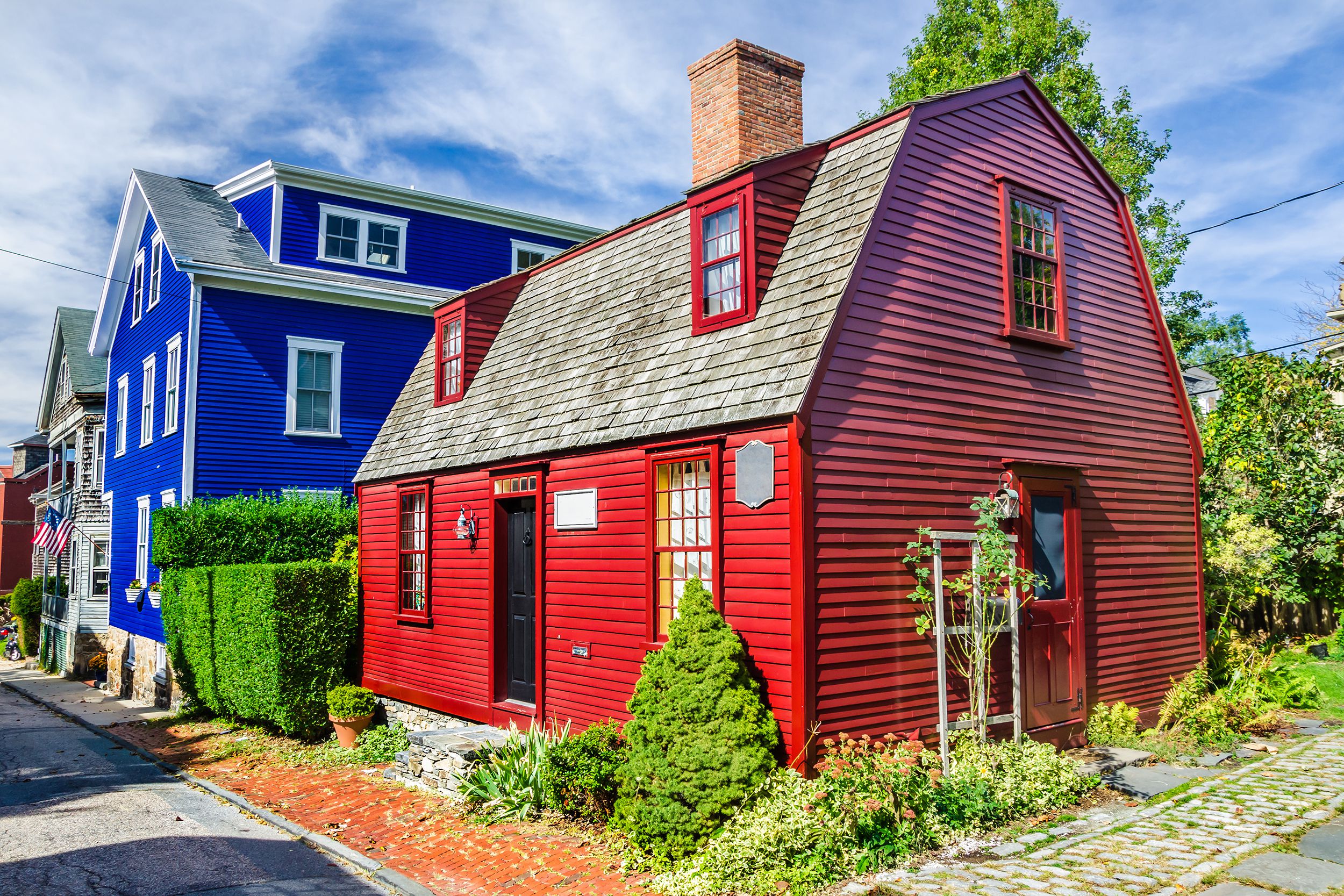 Red historic house in Newport, Rhode Island, in the foreground, a line of more historic houses to the left, one is blue, with small manicured yards, on a sunny day during summer