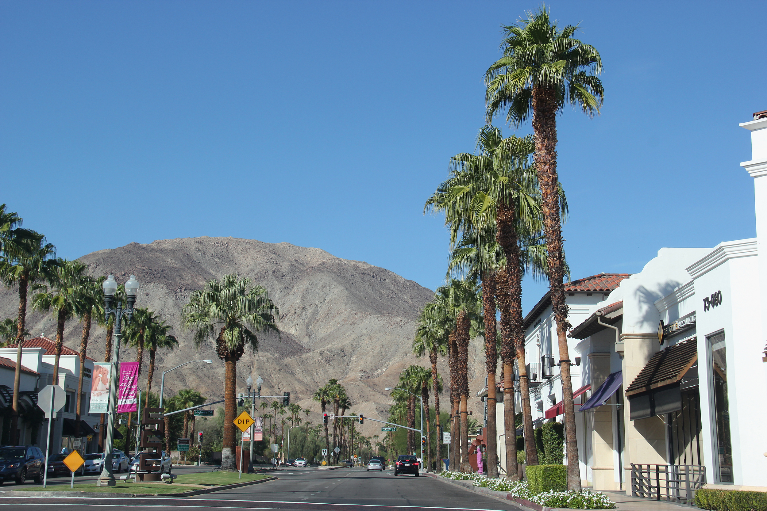 Road going through El Paseo Shopping District, Palm Desert, California, shopping on both sides with palm trees, on a sunny day with a large mountain in the background