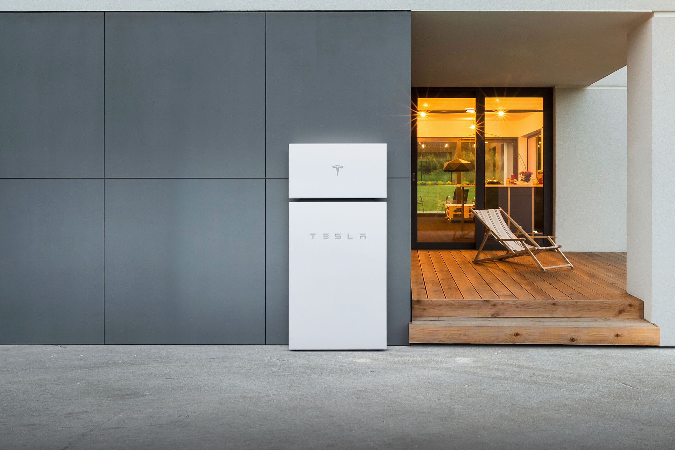 Tesla Powerwall along grey external wall of home, adjacent to porch and lit up inside living space