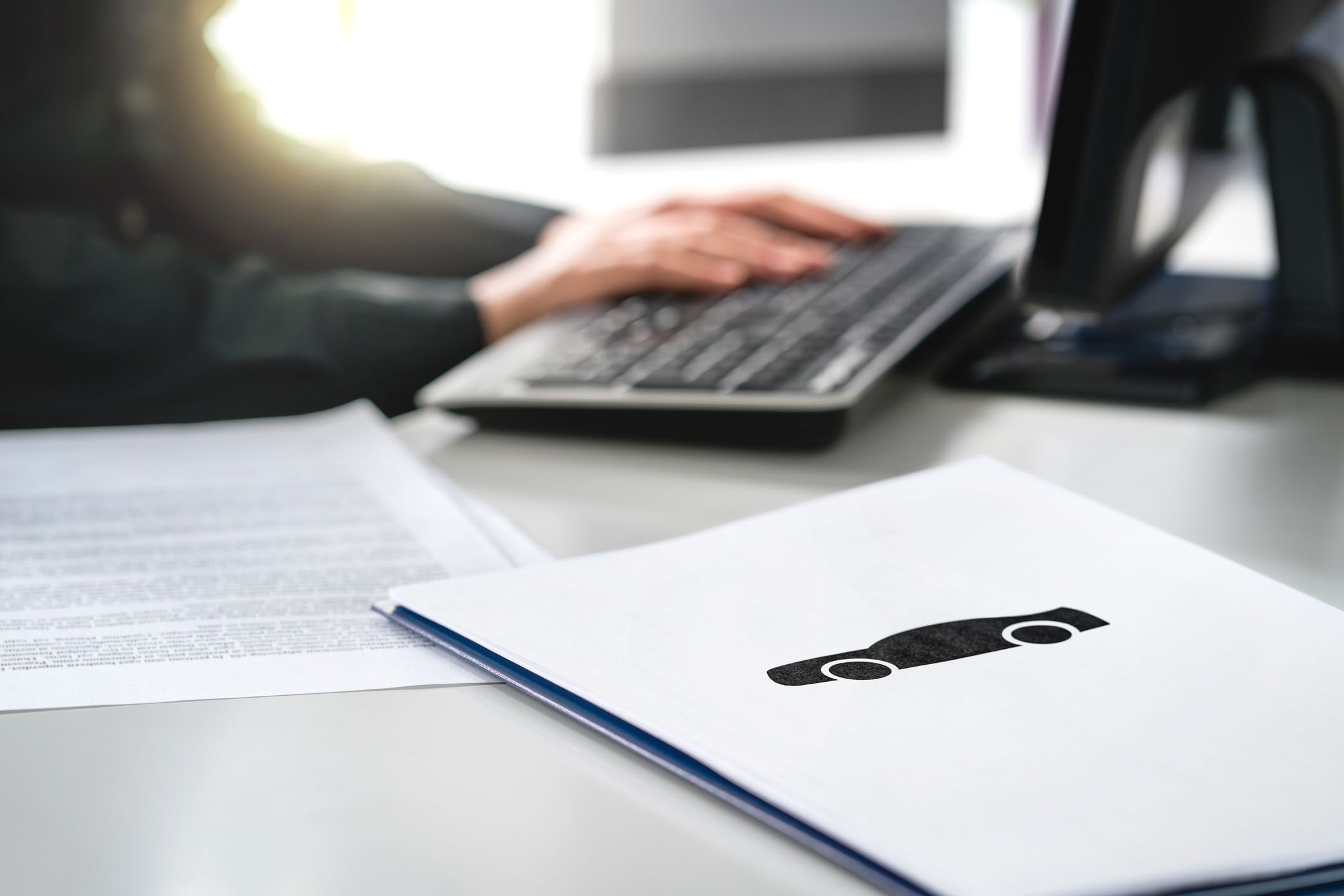 folder with car emblem in foreground, documents and hands on computer keyboard in background