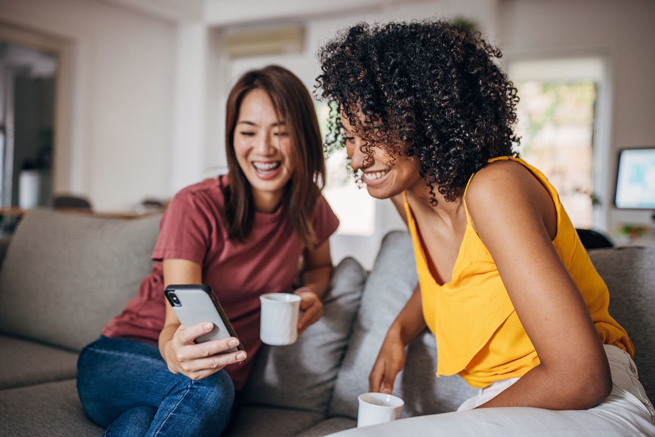Two people, diverse women sitting on sofa at home, they are using smart phone together.