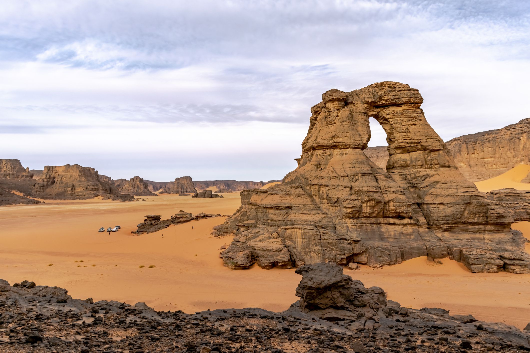 Far tiny tourists and 4x4 cars parked down. Arch rock natural window shape, mountain, orange color sand and blue cloudy sky. Tassili N'Ajjer park.