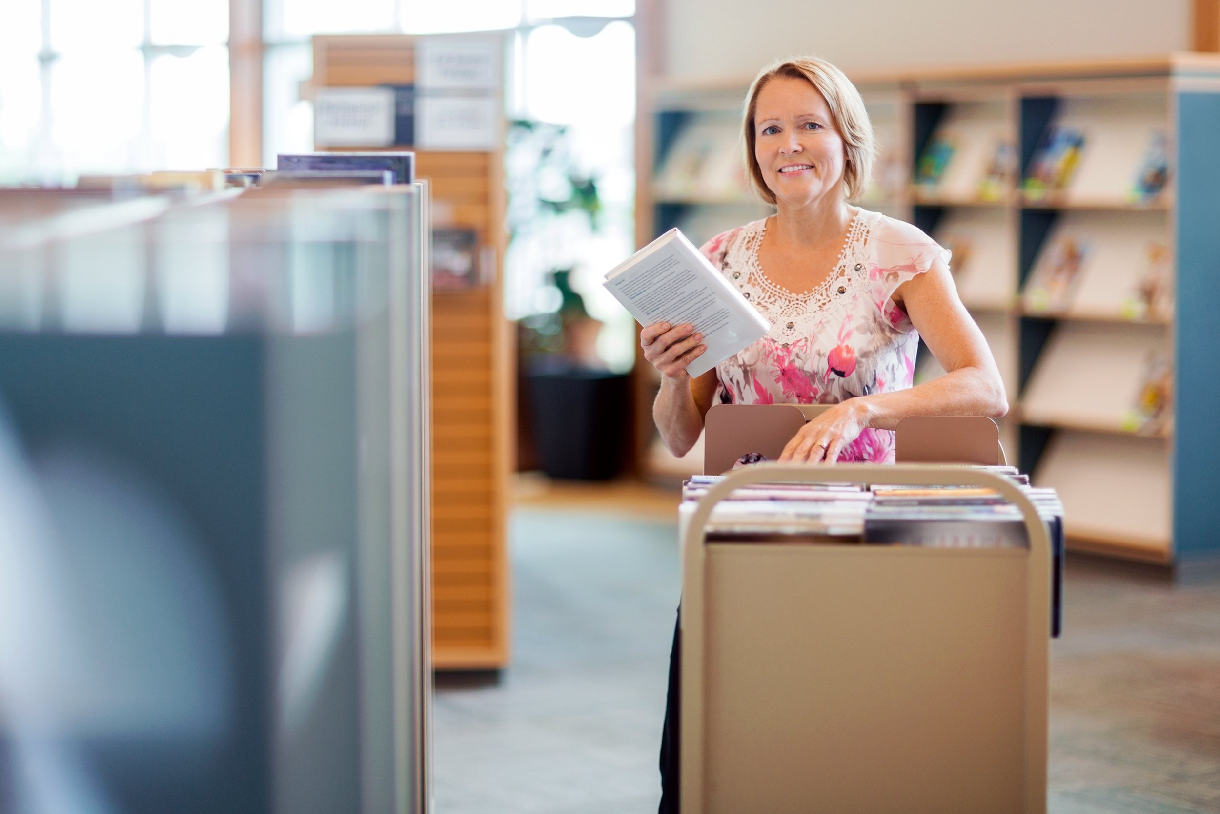 Portrait of smiling female librarian with trolley of books in library