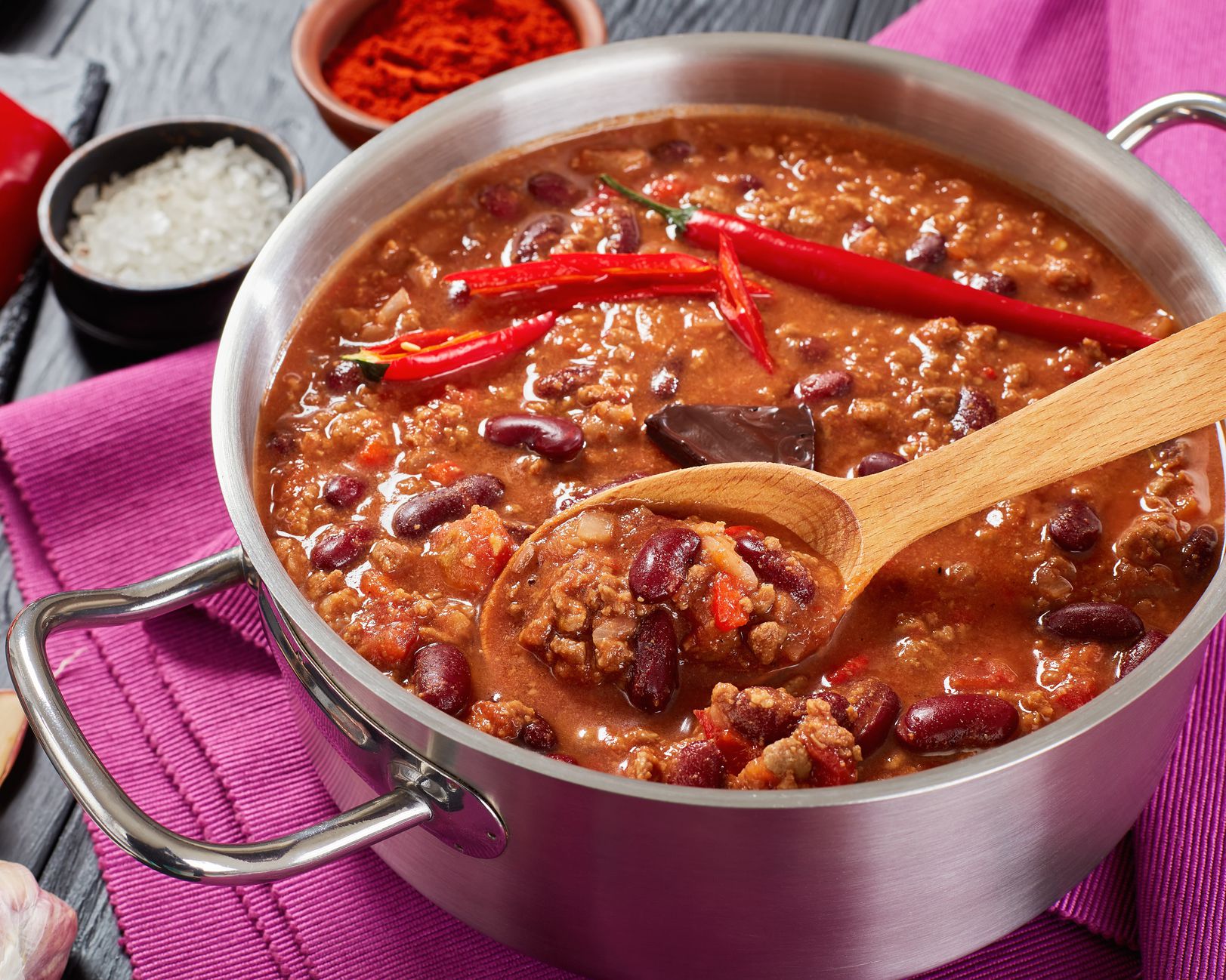 5-ingredient chili in a two-handled pot with a wooden serving spoon on a mauve napkin and dark grey wooden table with ingredients in the background