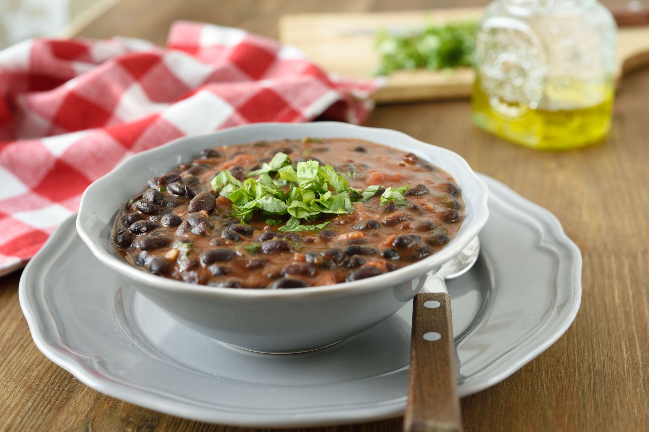 5 ingredient instant pot black bean soup in a white bowl on a matching decorative white plate with a spoon on a wooden table with a blurred background of ingredients