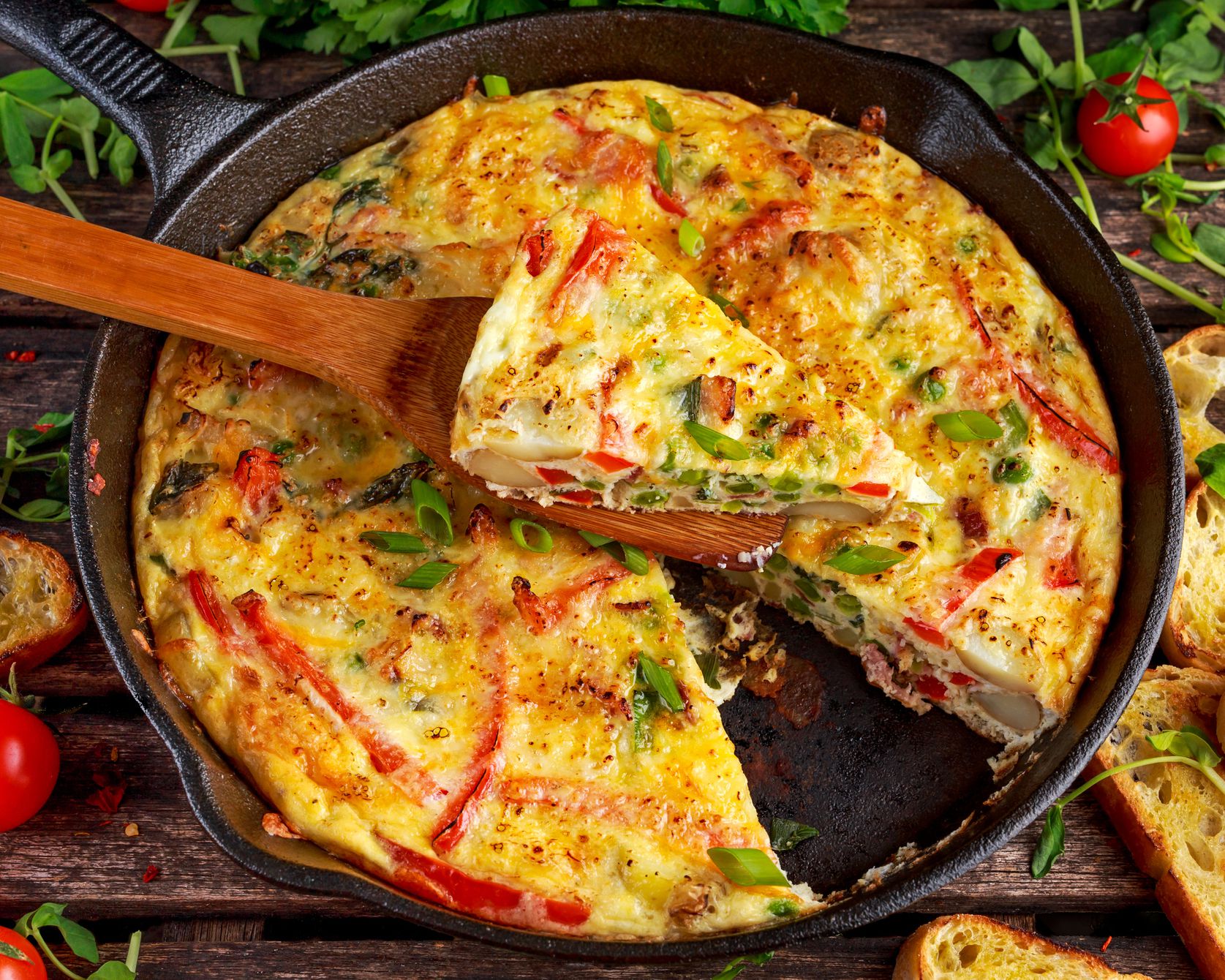 5 ingredient pressure cooker cheesy egg bake in a cast iron skillet with a wooden server holding one piece with a background of a wooden table, tomatoes, and pieces of garlic bread