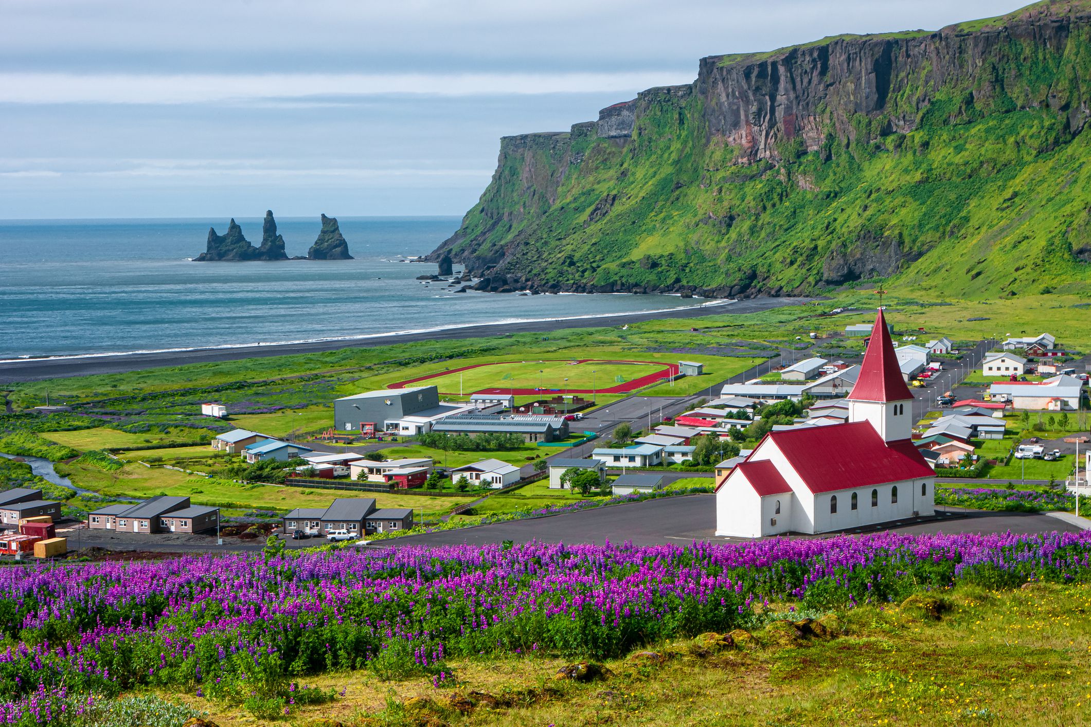 Basalt stacks and black sand beach of Reynisdrangar, South Iceland, vibrant green during summer, a small town and church