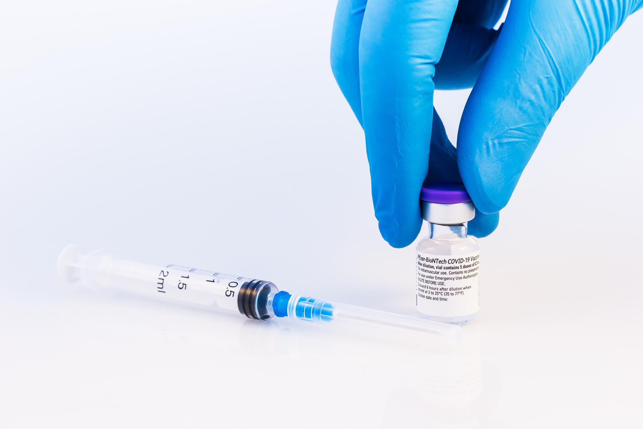 Blue gloved hand holding a vial of the Covid-19 vaccine on a white table, with a plastic syringe to the left of it, with a white background
