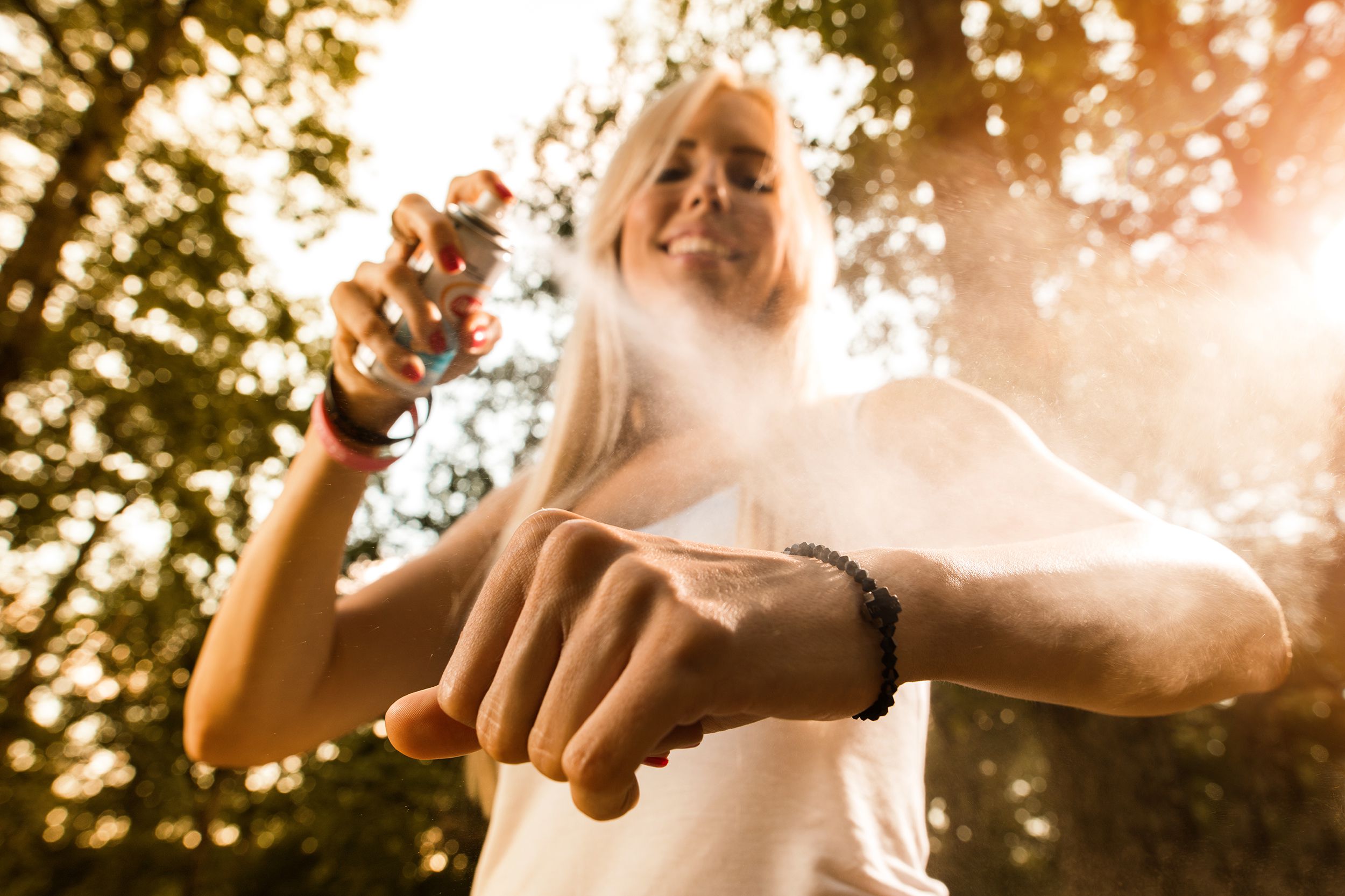 Close up of smiling woman applying insect repellant on her arm, dramatic angle with hand and arm in the foreground, surrounded by woods outside
